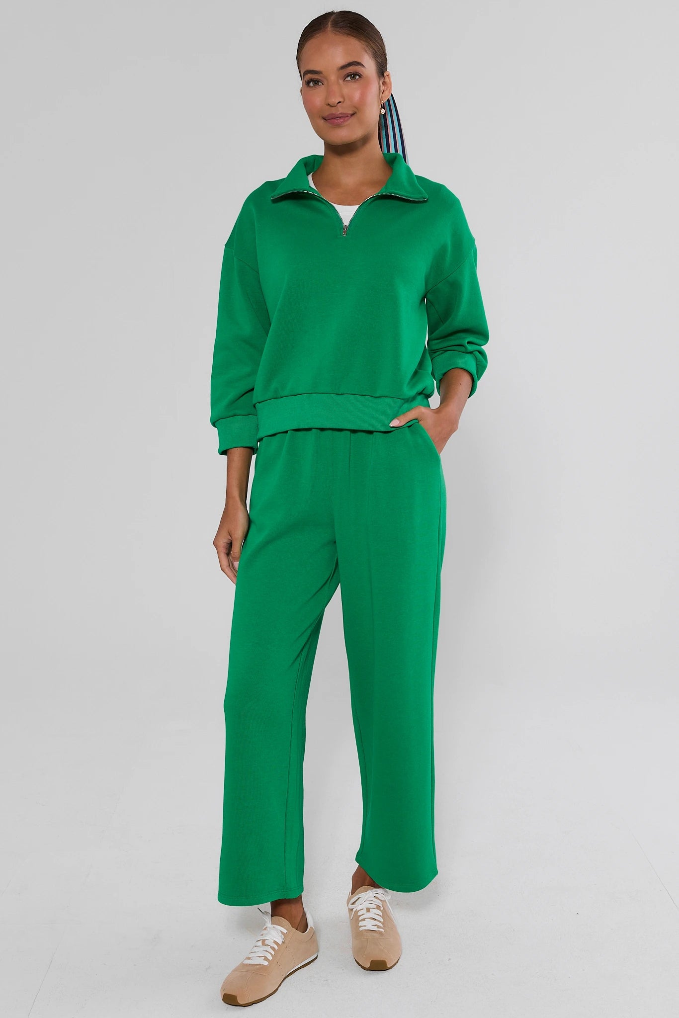 A woman modeling a bright green two-piece tracksuit consisting of a long-sleeve half-zip pullover and matching wide-leg pants, paired with beige sneakers, against a light gray studio background.
