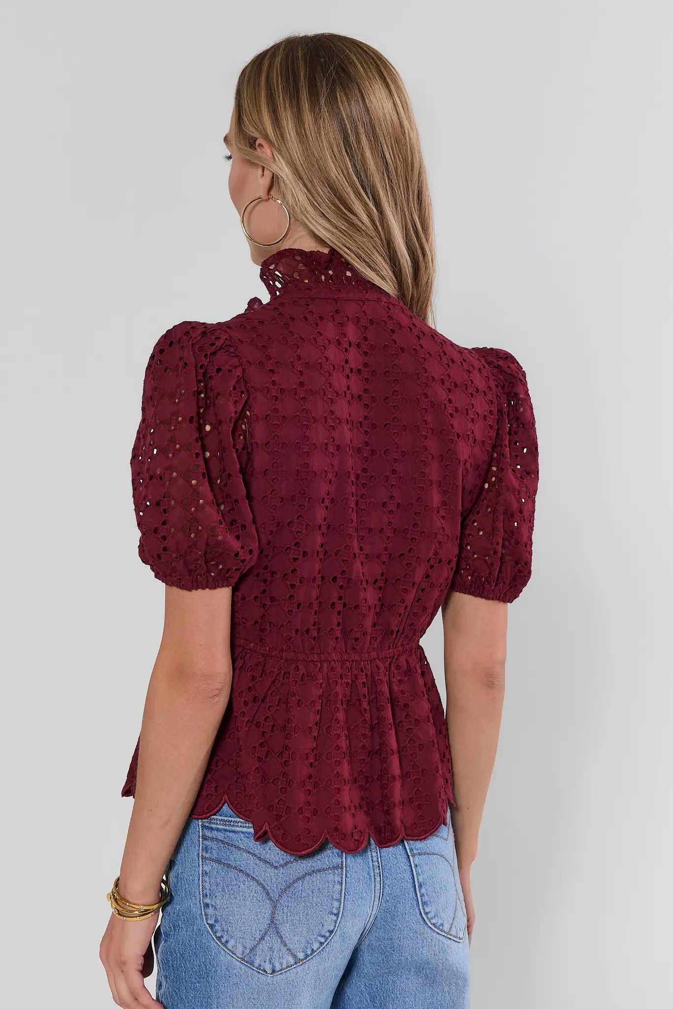 A woman viewed from the back wearing a burgundy eyelet lace blouse with puff sleeves and a ruffled hem, paired with light blue jeans against a plain light gray background.