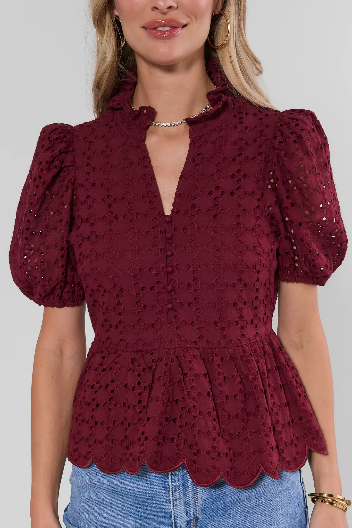 A woman wearing a burgundy eyelet lace top with puff sleeves, a split neckline, and a scalloped hem, paired with blue jeans against a light gray background.