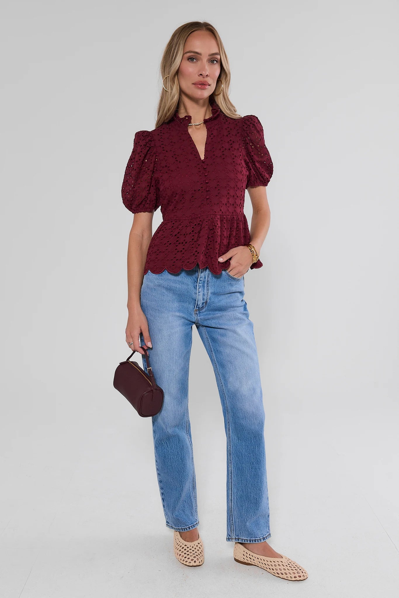 A woman modeling a burgundy short-sleeve lace top with puff sleeves and a scalloped hem, paired with light blue jeans, a small dark burgundy handbag, and beige woven flat shoes against a light gray studio background.