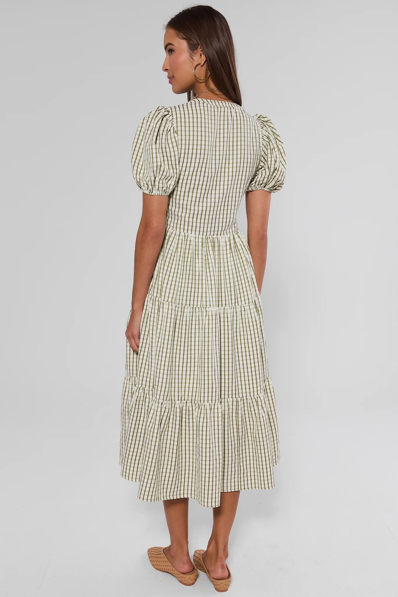 A woman shown from the back wearing a cream and green gingham tiered midi dress with puff sleeves, paired with tan flat sandals, against a light gray studio background.