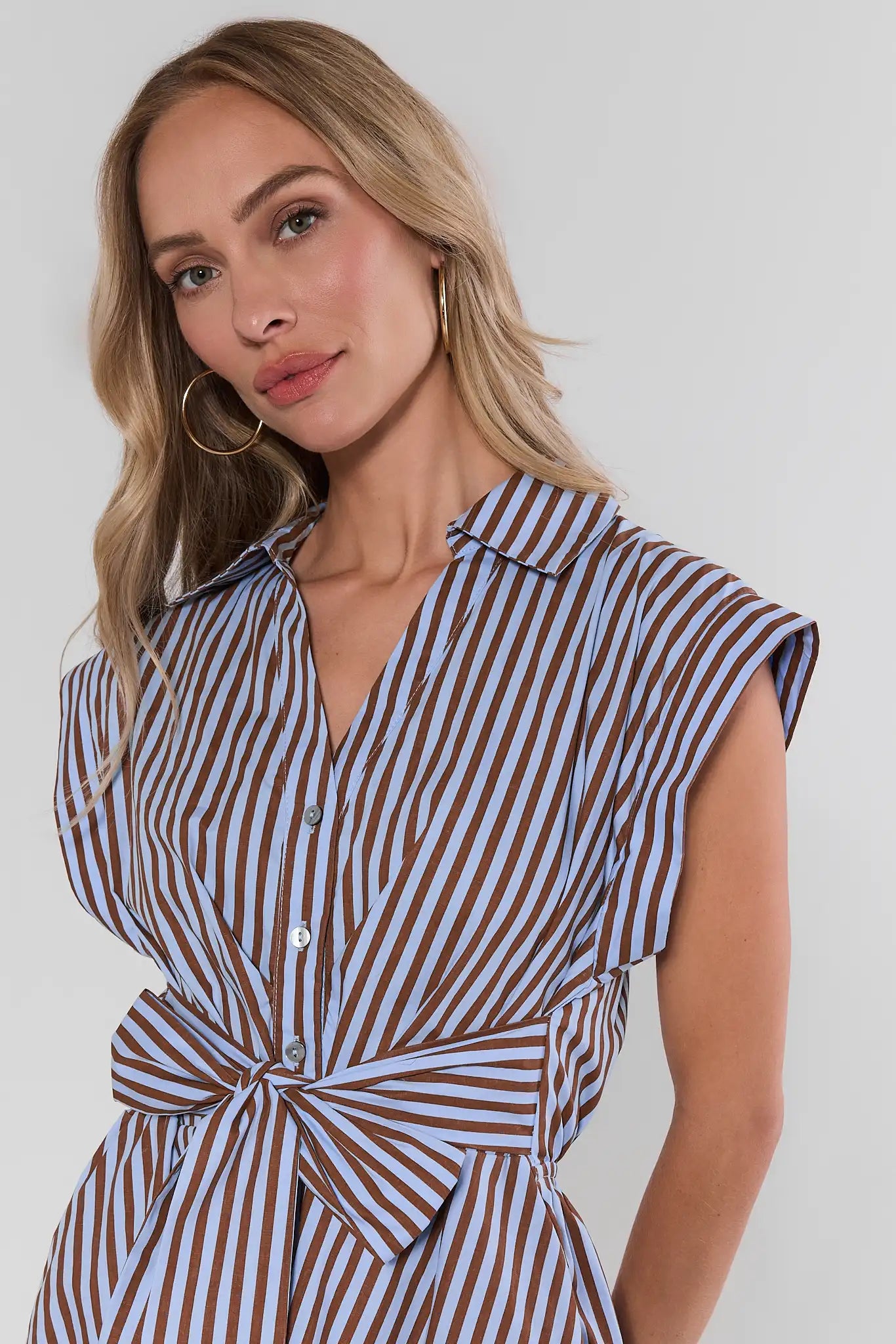 A woman wearing a brown and light blue vertically striped shirt dress with a tie waist, shown from the chest up against a plain light gray background.