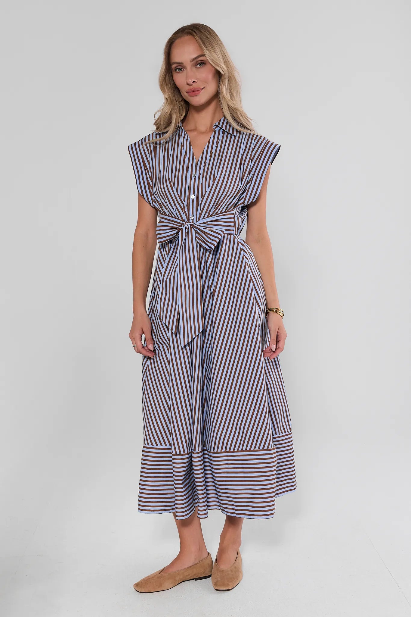 A woman modeling a sleeveless midi dress with vertical navy, white, and red stripes, featuring a tie waist and button collar, set against a plain light background.