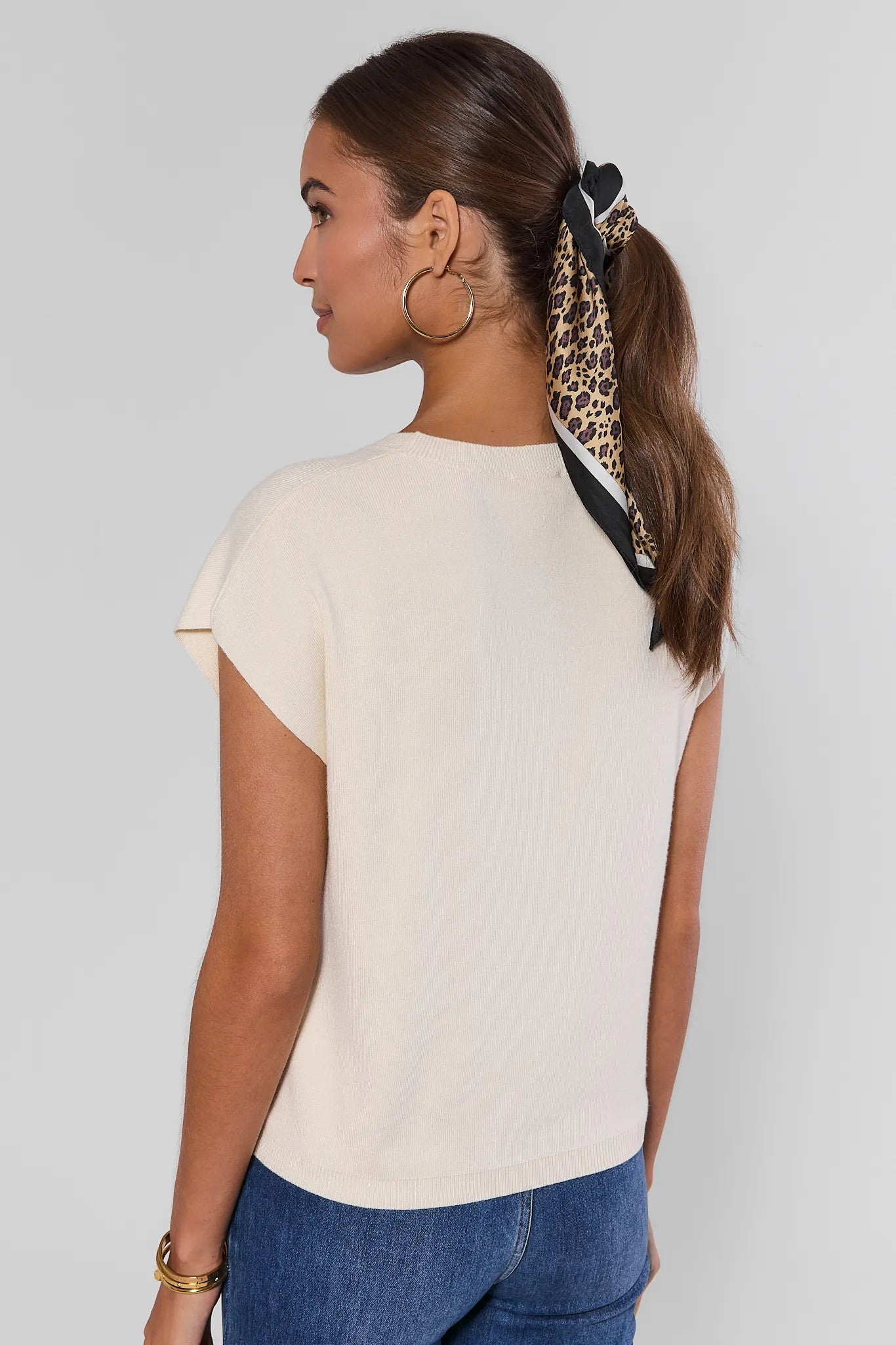 A woman shown from the back wearing a short-sleeve cream-colored top and blue jeans, with her hair tied back in a ponytail using a leopard-print and black hair tie, against a light gray studio background.