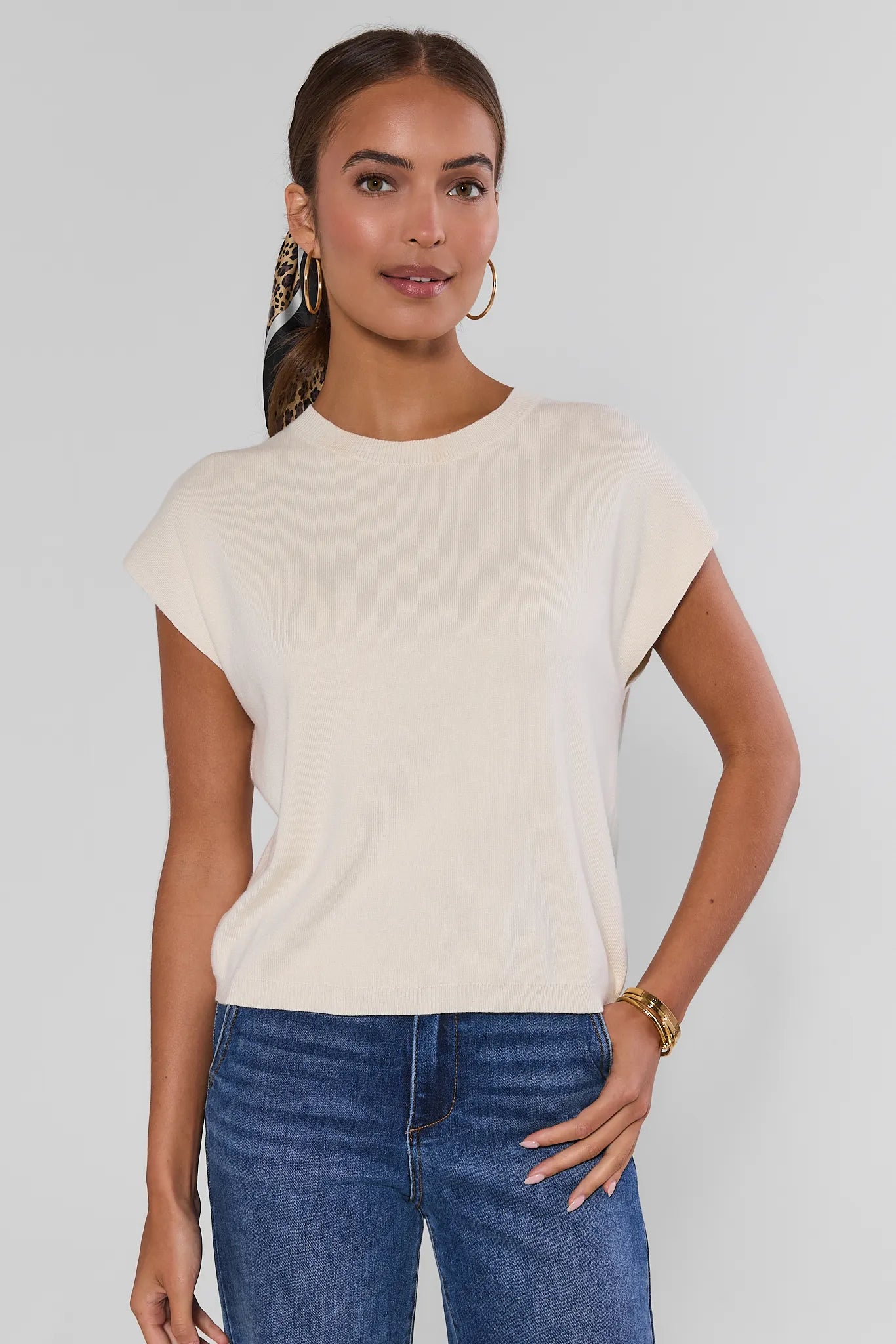 A woman modeling a short-sleeve crewneck top in a light cream color, paired with blue jeans, against a plain light gray background.