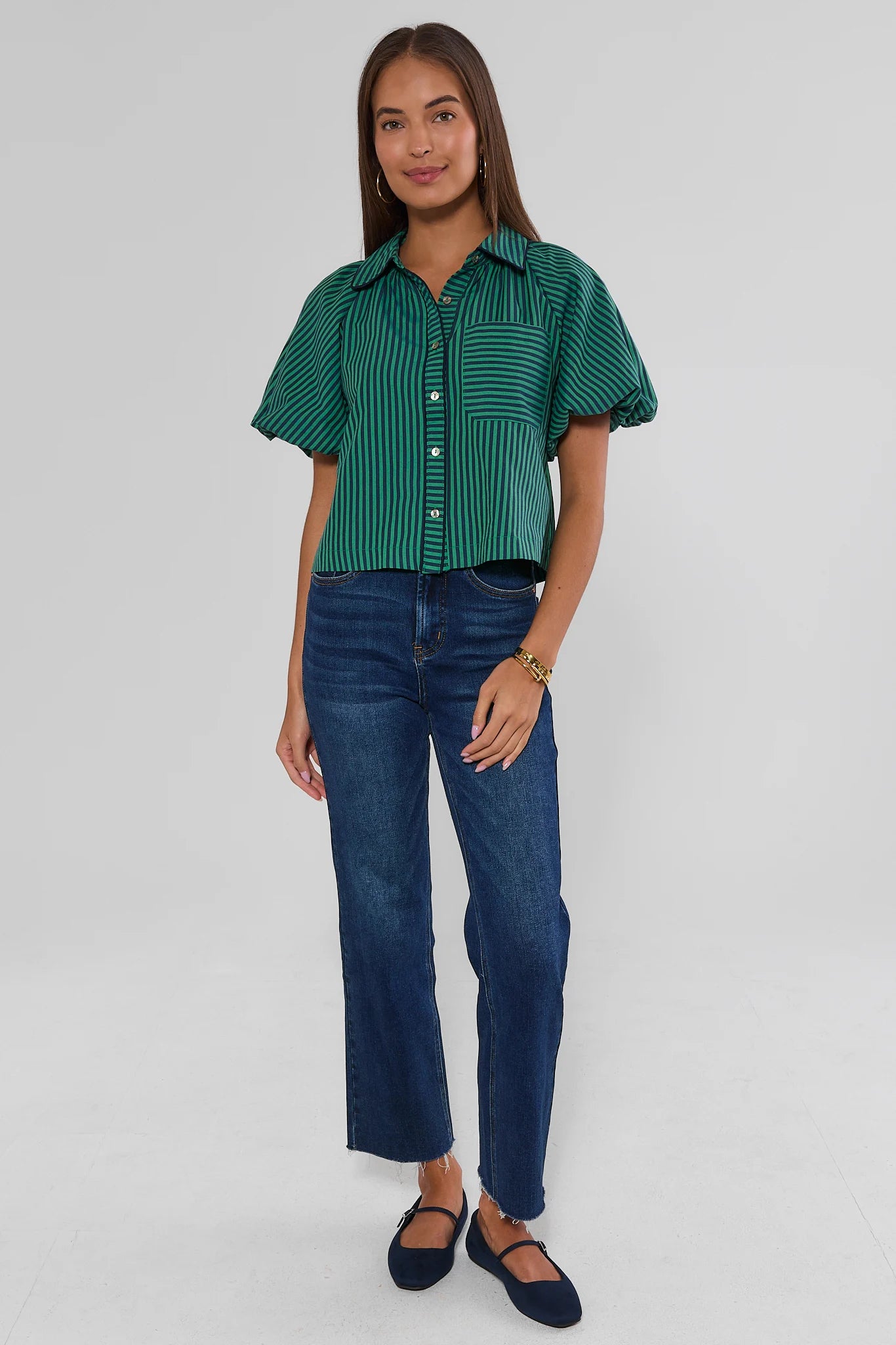 A woman modeling a green and white vertically striped short-sleeve button-up shirt paired with dark blue jeans and navy flats, standing against a light gray studio background.