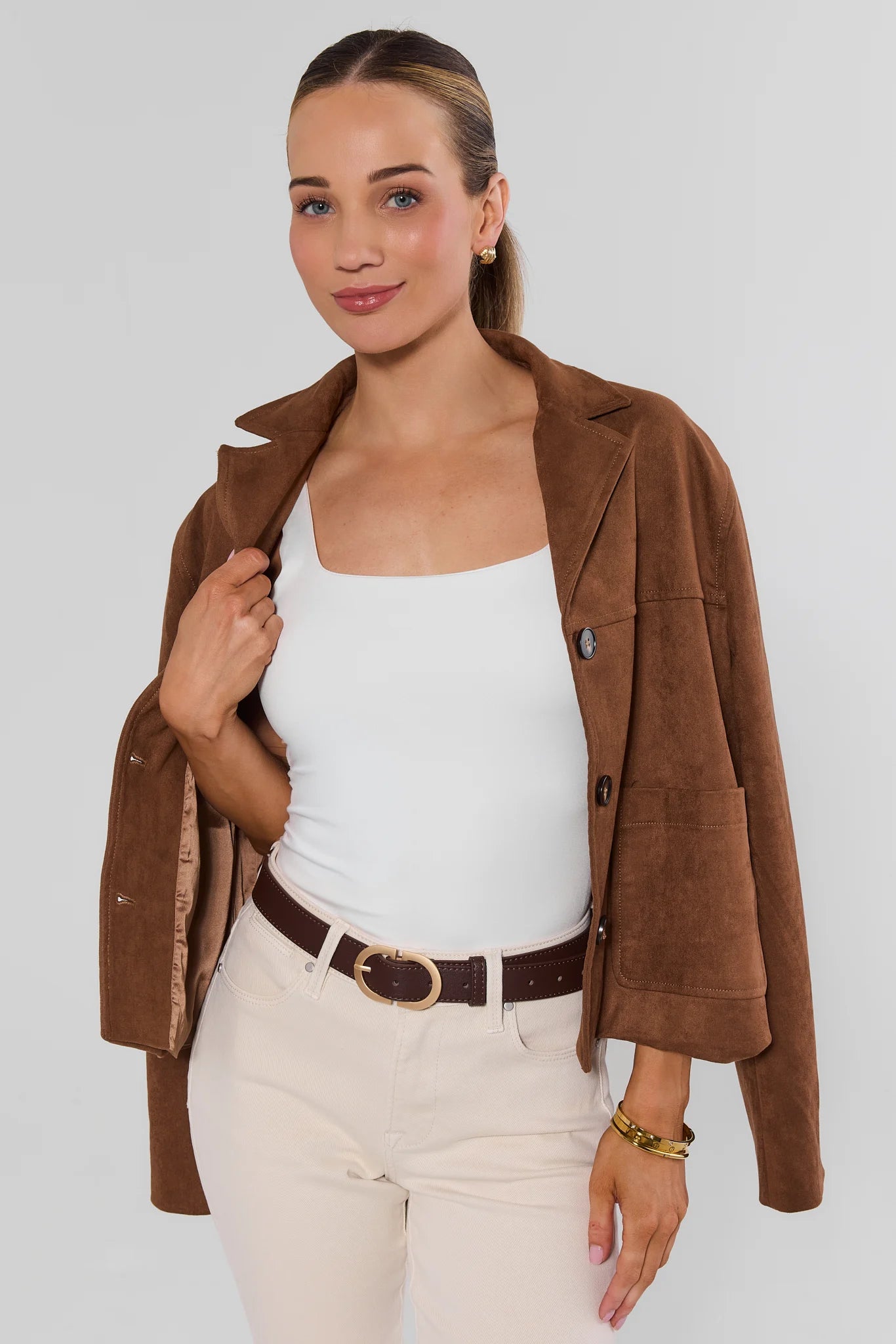 A woman modeling a brown suede-like blazer over a white tank top and cream pants, standing against a light gray studio background.