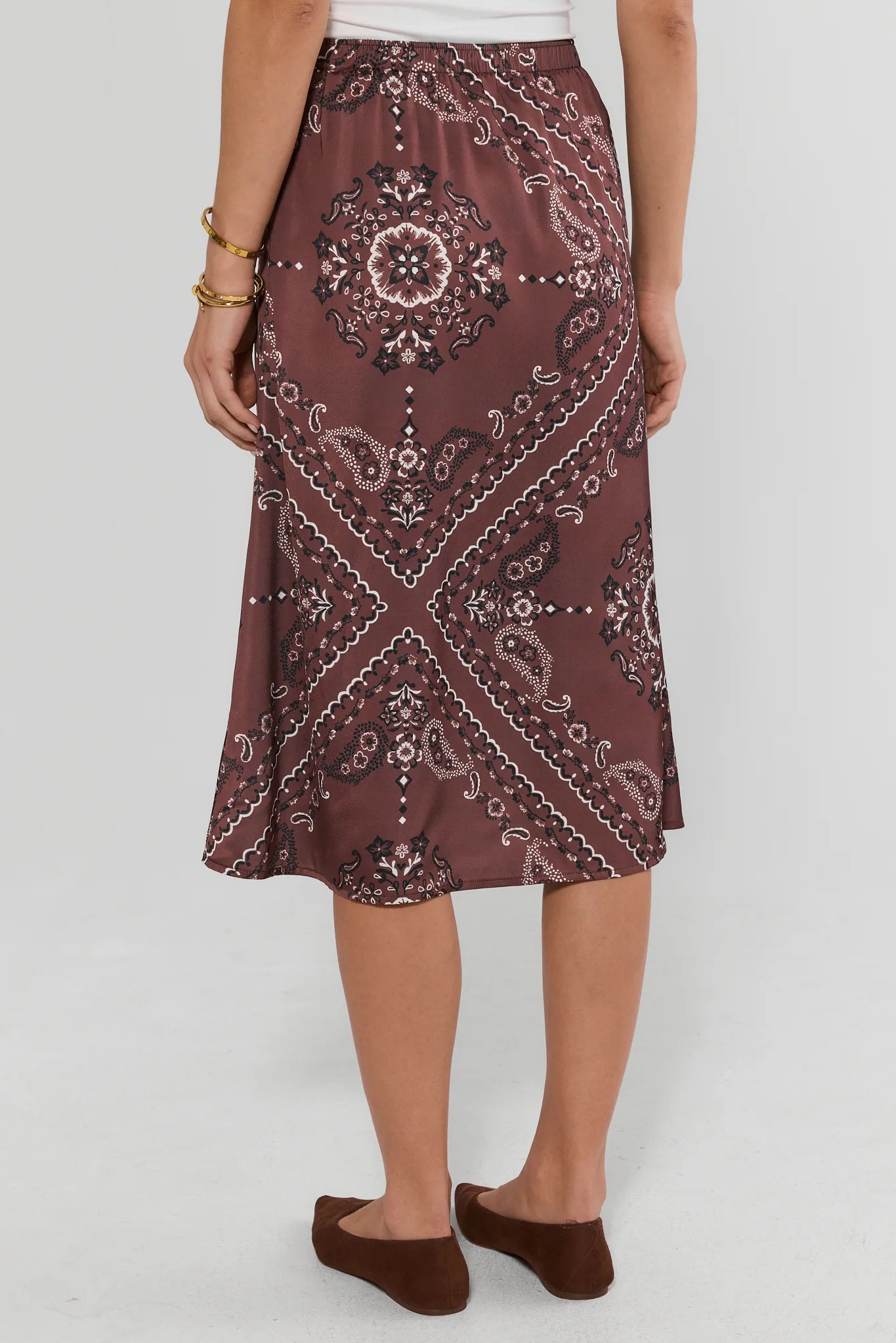 A woman wearing the Brown Bandana Print Midi Skirt with a white and black paisley bandana pattern, paired with brown slip-on shoes against a light gray studio background.