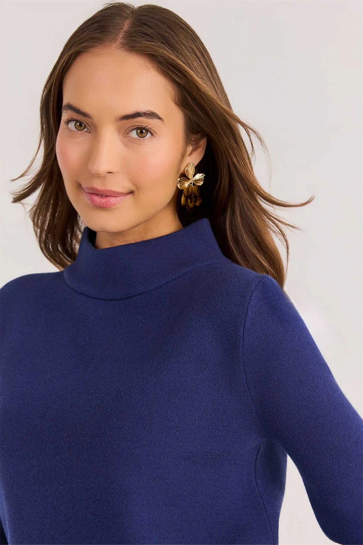 A woman with long brown hair wearing a navy blue mock neck sweater and gold butterfly earrings, shown in a close-up portrait against a light gray background.