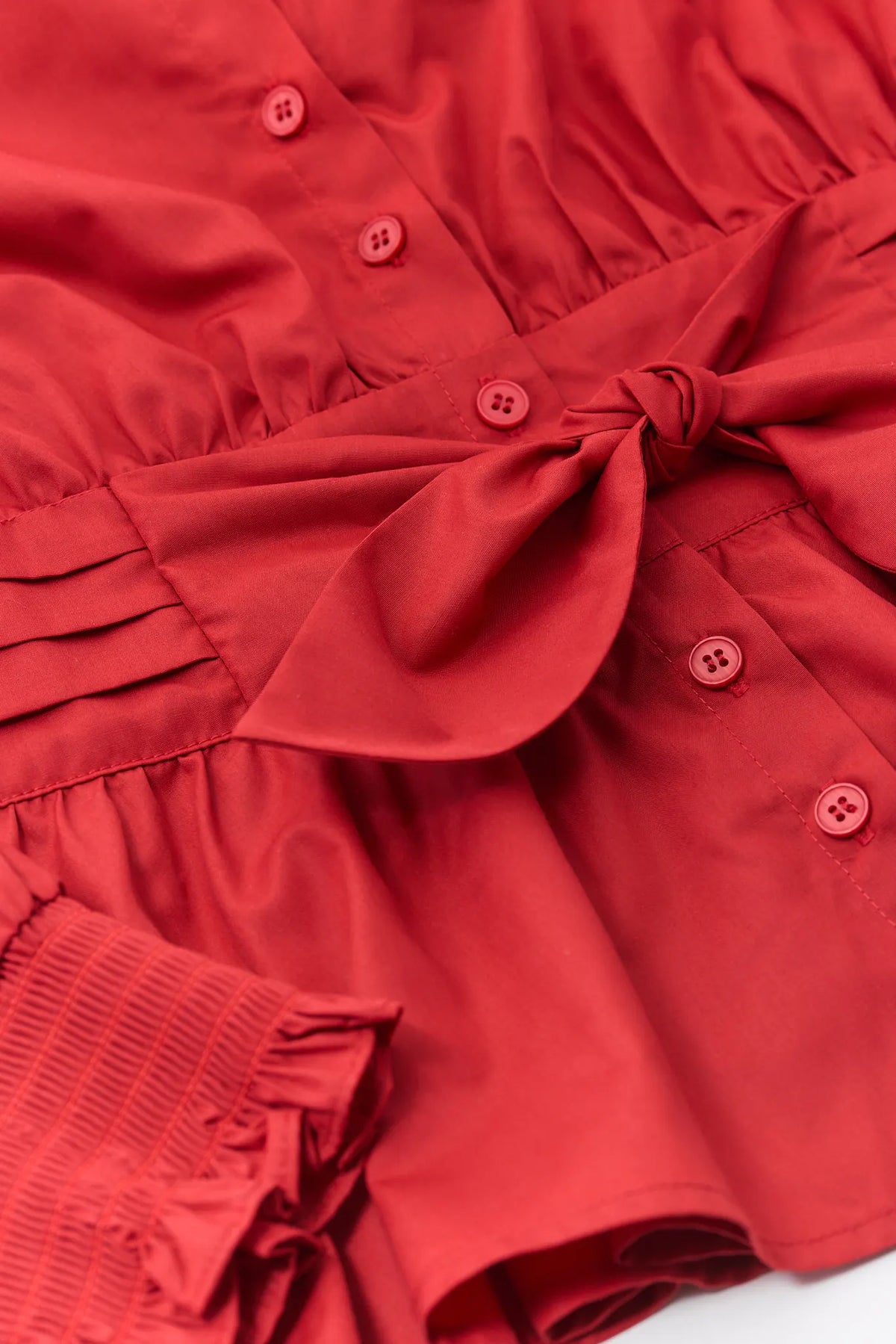 A close-up of a red fabric garment featuring a matching fabric tie belt, pleated detailing, and small matching buttons.