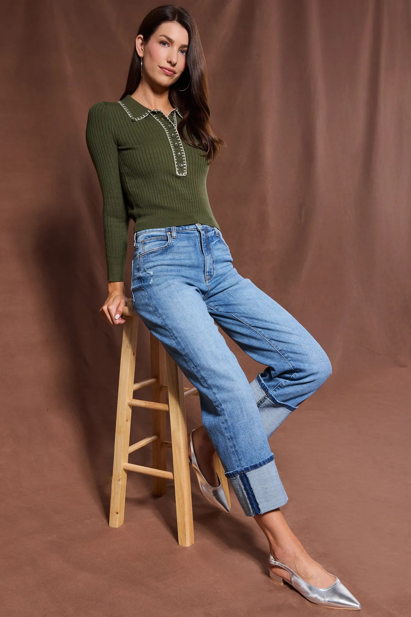 Woman wearing a green Shayla Sweater and blue jeans sitting on a wooden stool against a brown background