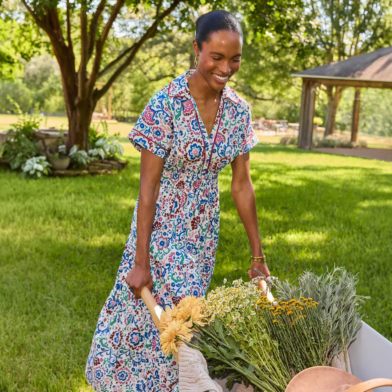 A woman stands outdoors in a grassy yard wearing a short-sleeve collared dress with a colorful floral and medallion print. She is smiling and holding a basket of freshly harvested flowers.