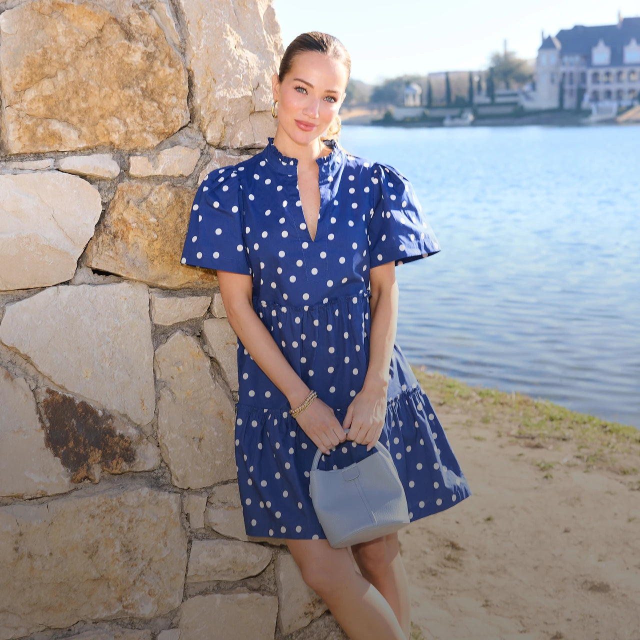 Woman in a blue polka dot dress standing by a stone wall with water in the background