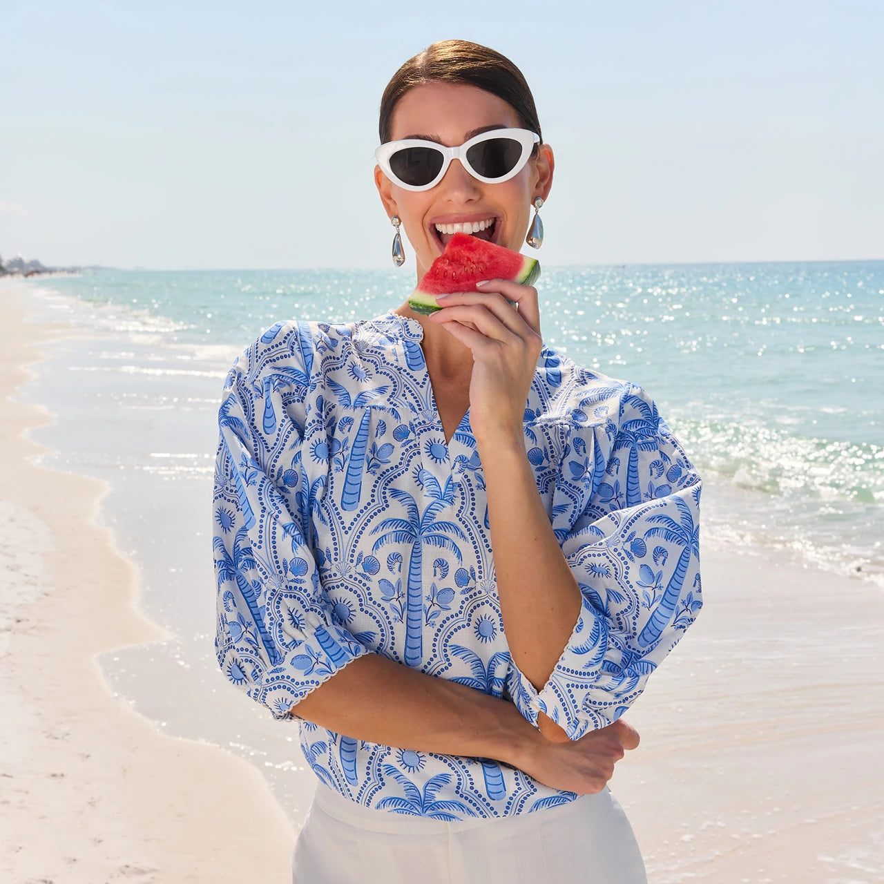 Woman in a blue and white patterned blouse eating watermelon on a beach. desktop