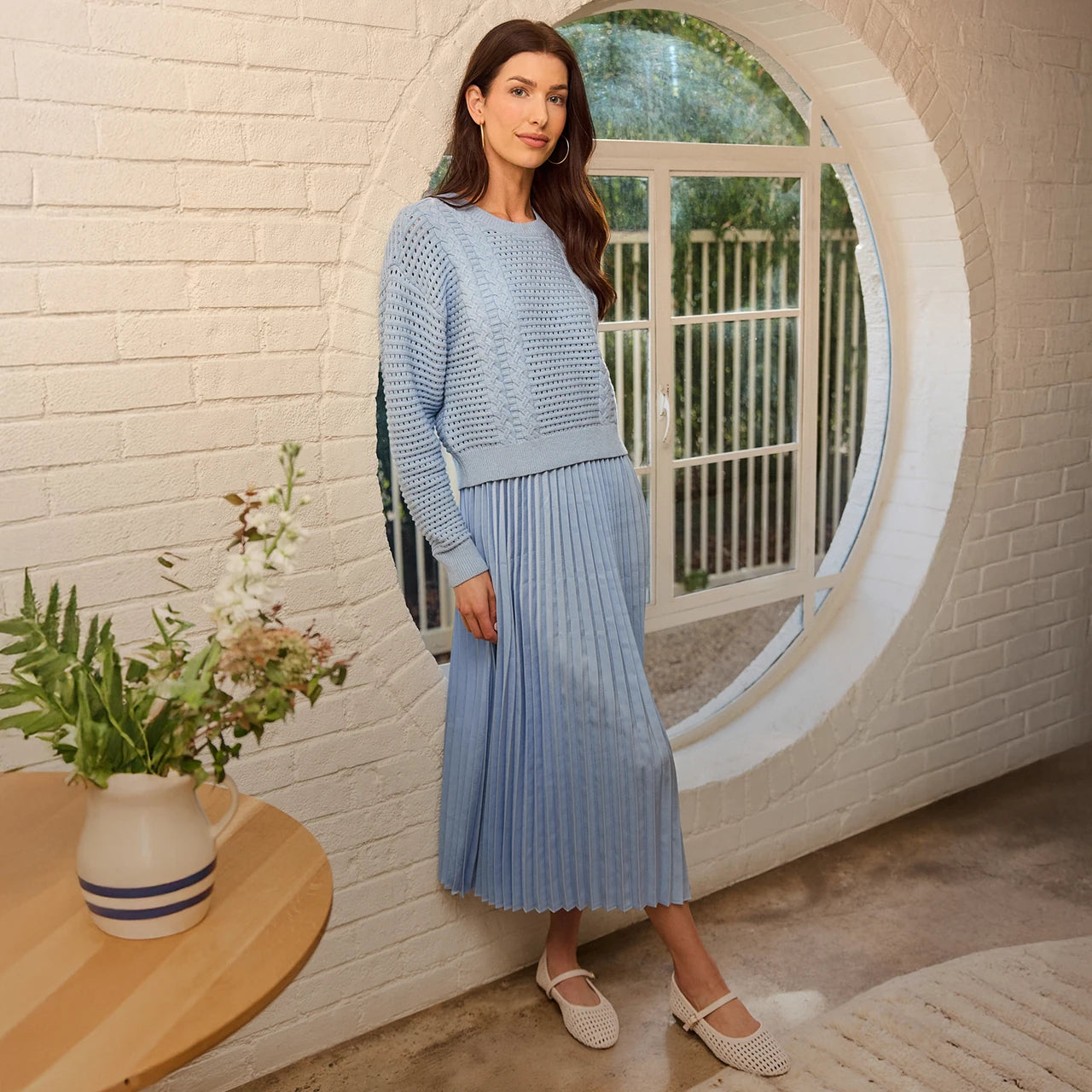 Woman in a light blue outfit standing in a room with a round window and a table with flowers.