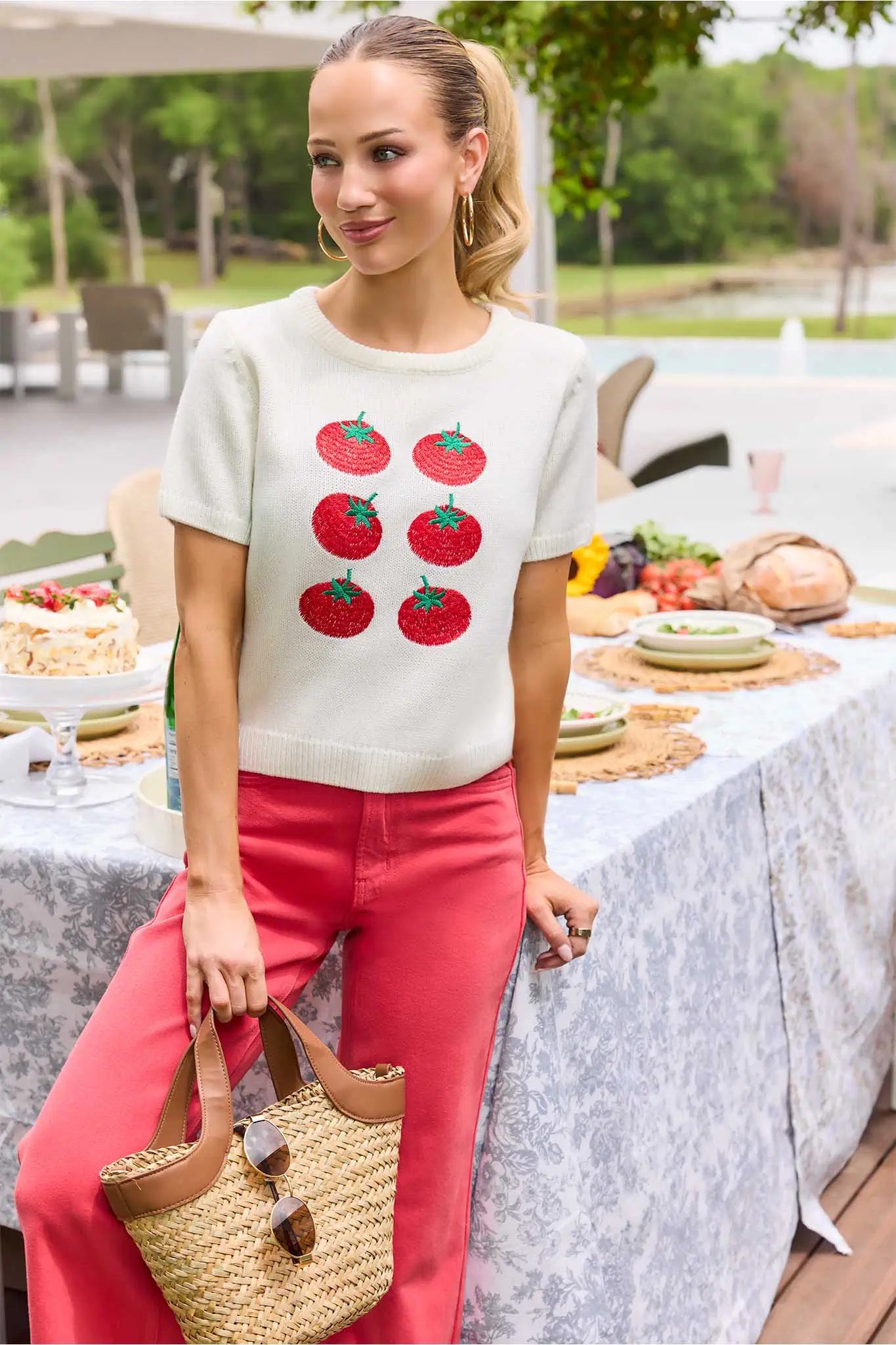 Woman in a tomato-patterned shirt and pink pants standing at an outdoor table setting.