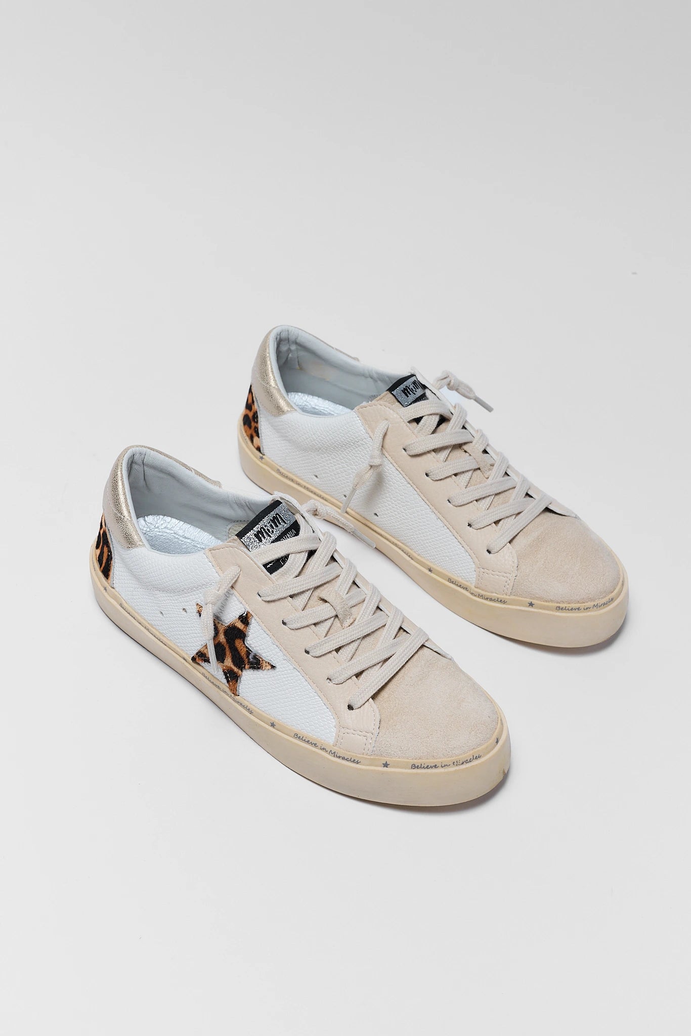 A pair of low-top sneakers in off-white suede and canvas, featuring a leopard-print star on the side and matching leopard-print heel accents, photographed on a light background.