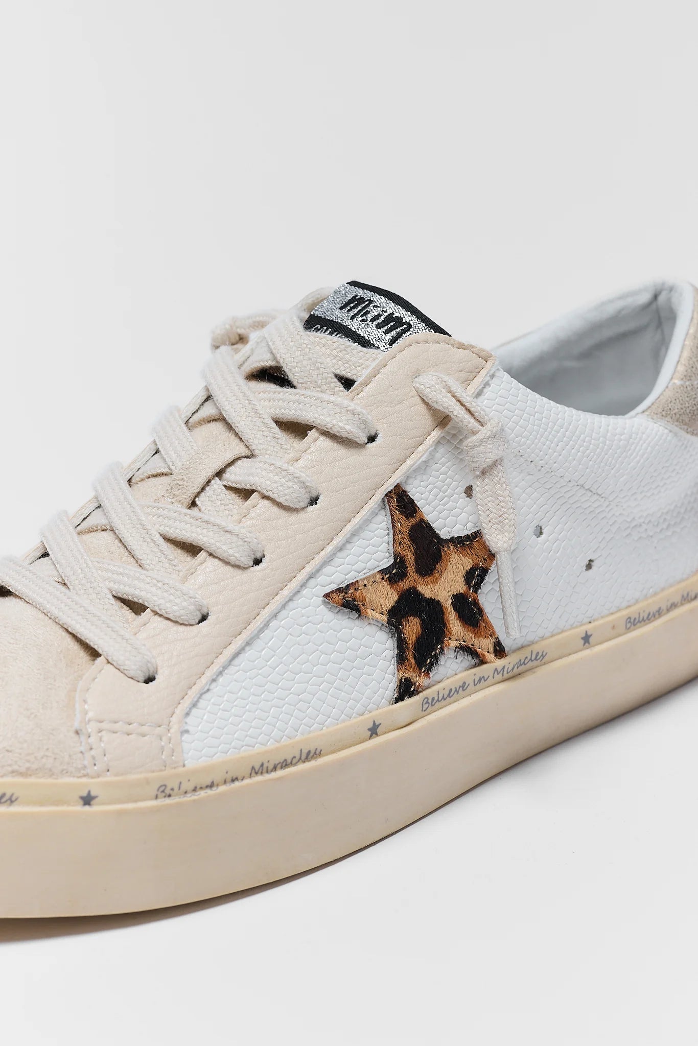 A close-up of a low-top sneaker in off-white and beige suede, featuring a textured white side panel, beige laces, a leopard-print star patch on the side, and a beige rubber sole.