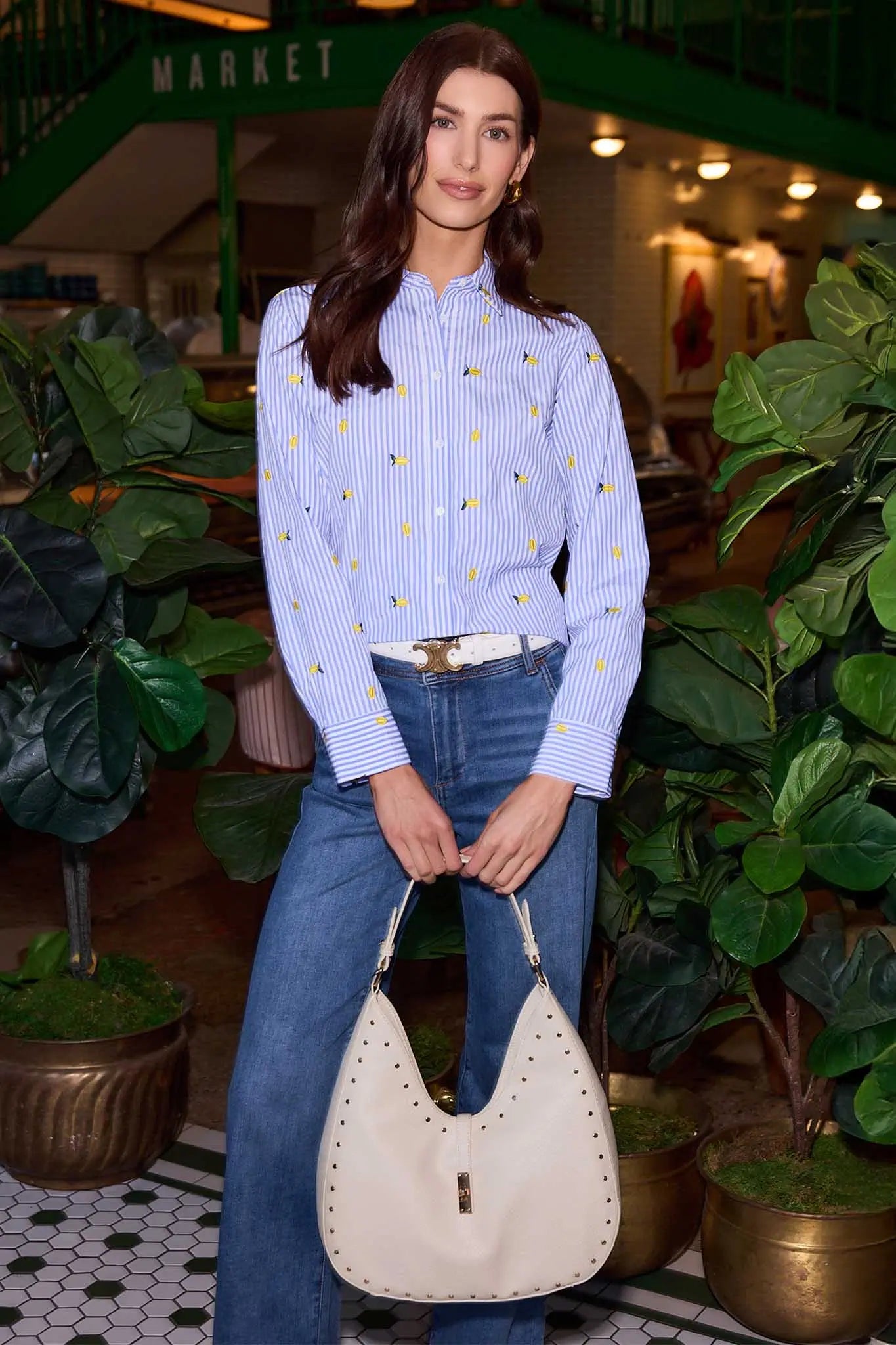 Woman holding a beige handbag in an indoor setting with plants and a market sign.