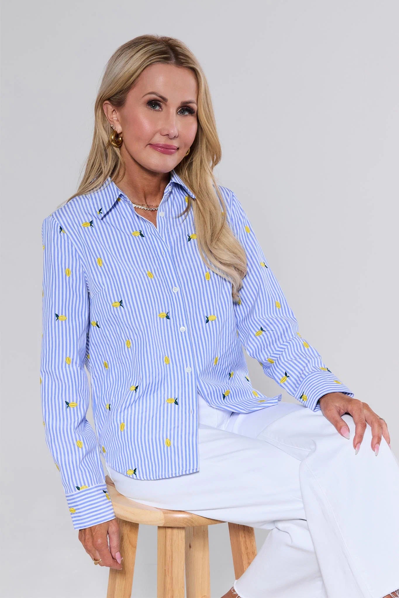 Woman wearing a blue striped shirt with yellow patterns and white pants, sitting on a wooden stool against a plain background.