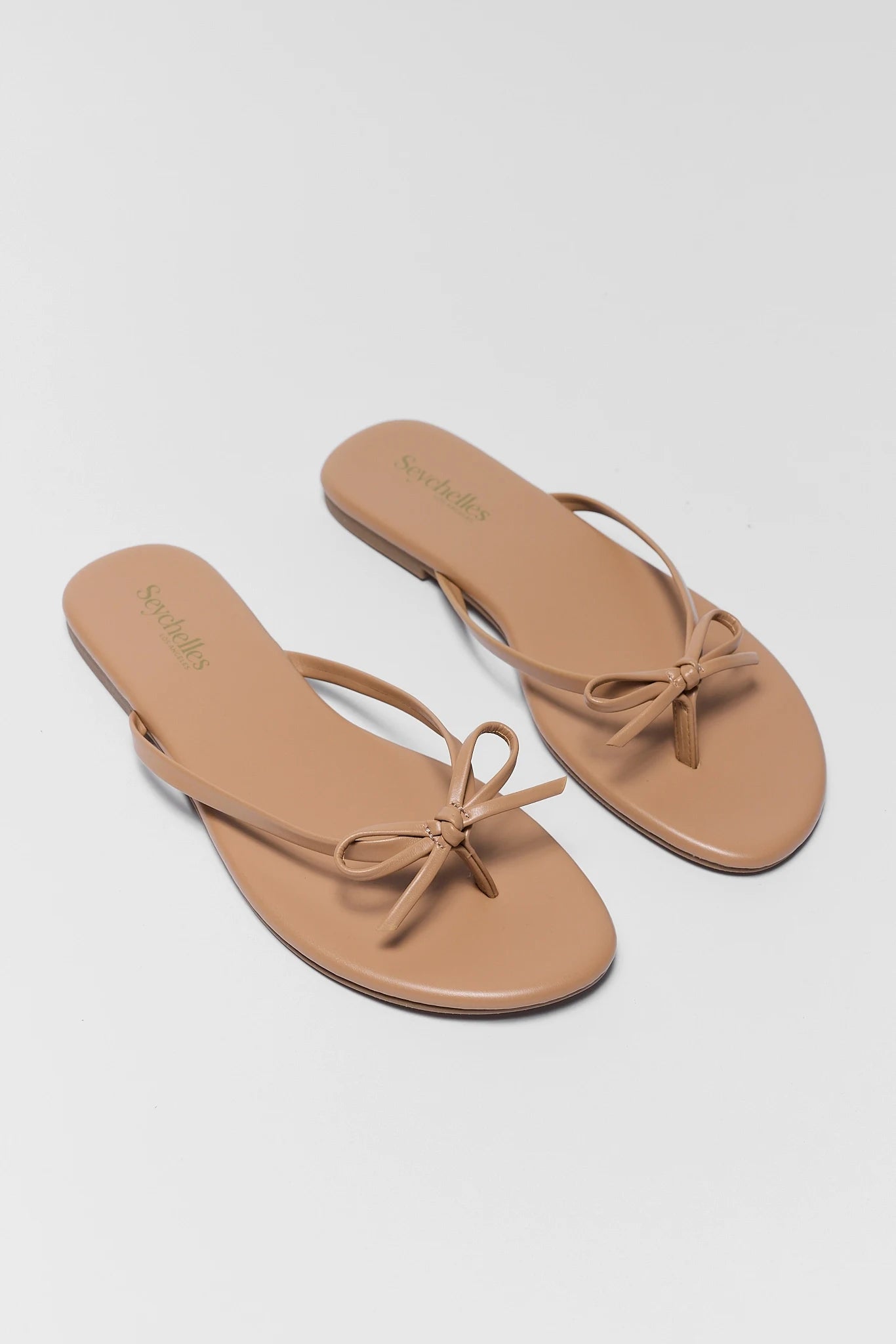 two Pair of tan sandals with bow detail on a light gray background