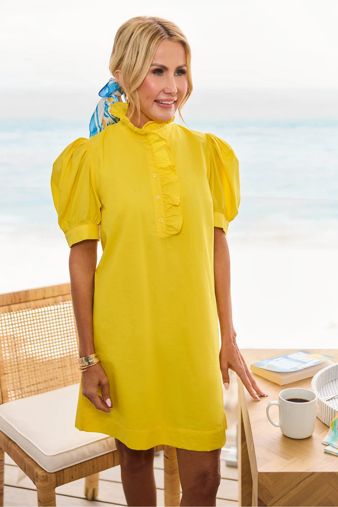 Woman in a yellow dress standing by a table with a coffee cup and notebook, with a blurred beach background.