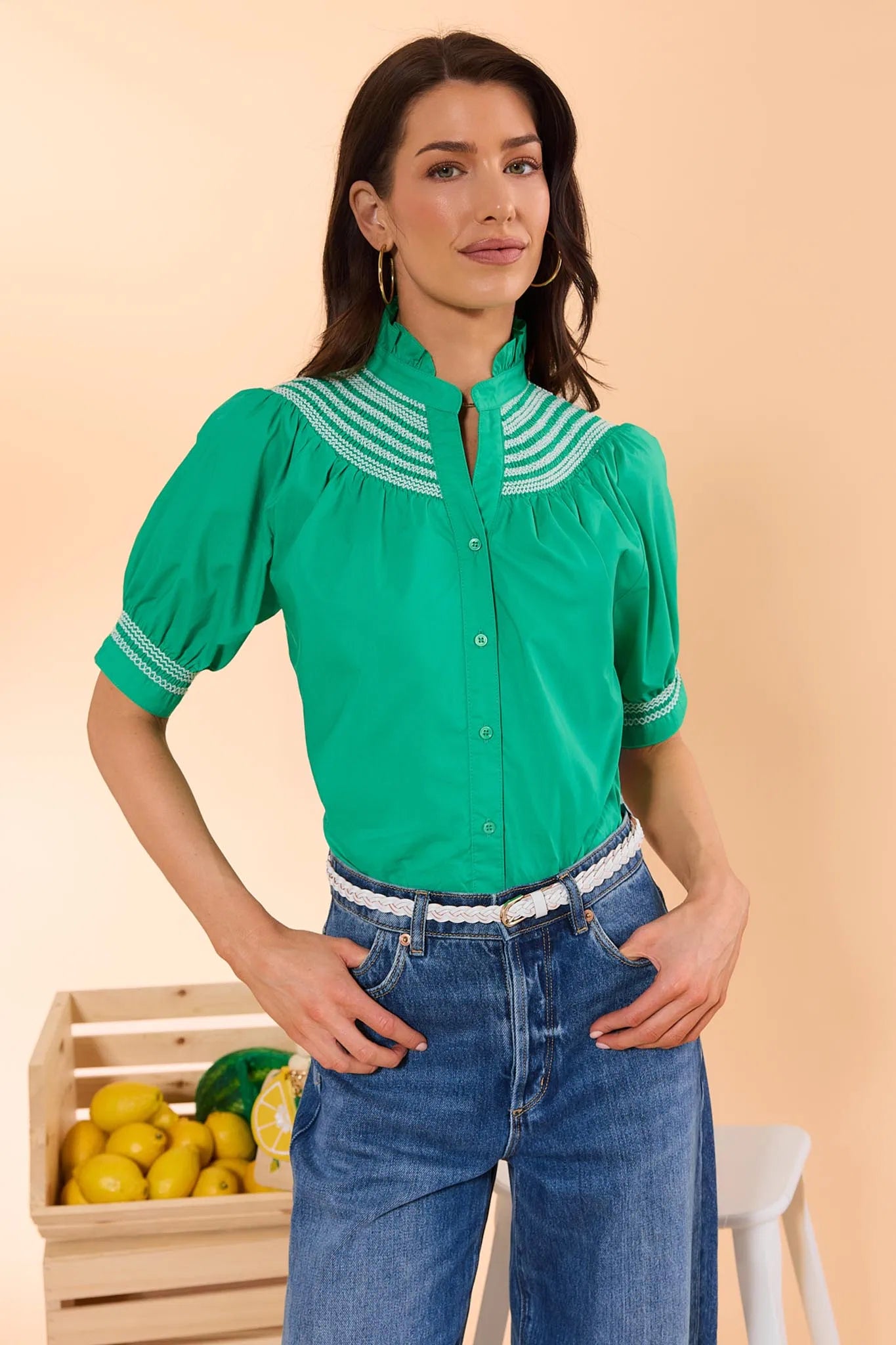Woman wearing a green blouse with white embroidery and blue jeans, standing next to a wooden crate of lemons on a beige background.