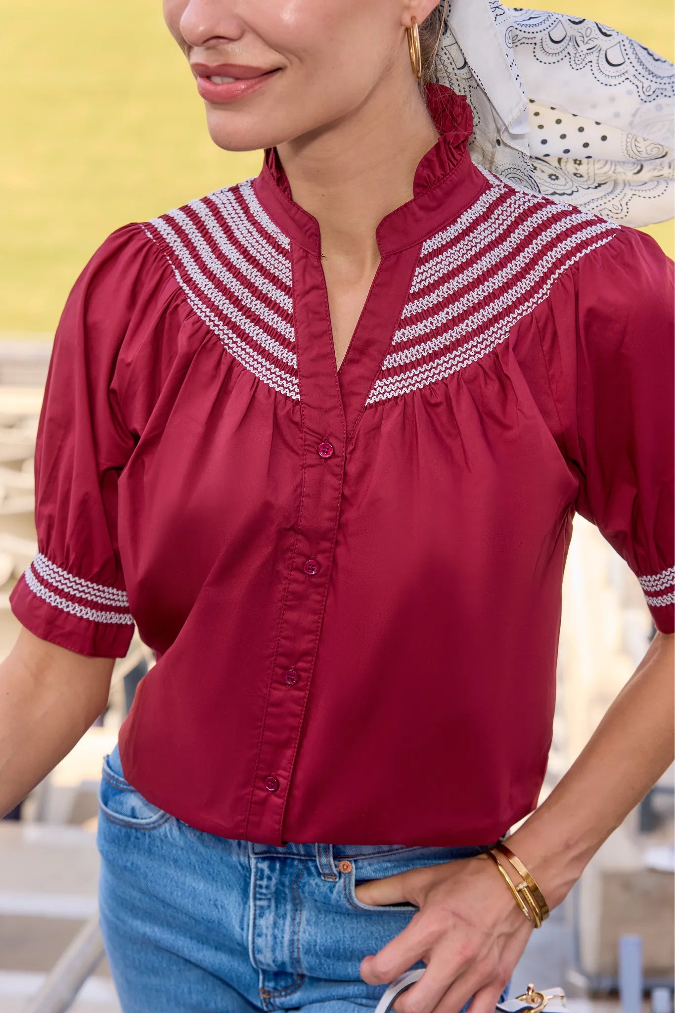 A woman wearing a short-sleeve button-front blouse in deep red with white embroidered stripes around the collar and cuffs, paired with blue jeans.