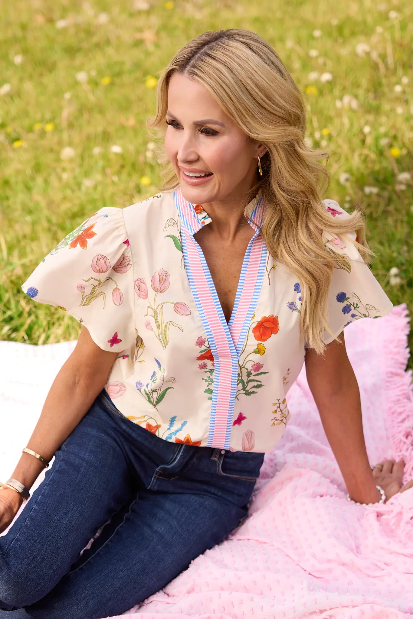 Woman in a Kieran Cream Floral Print Top, featuring a floral pattern and striped trim, sitting on a pink blanket with jeans in a grassy field.
