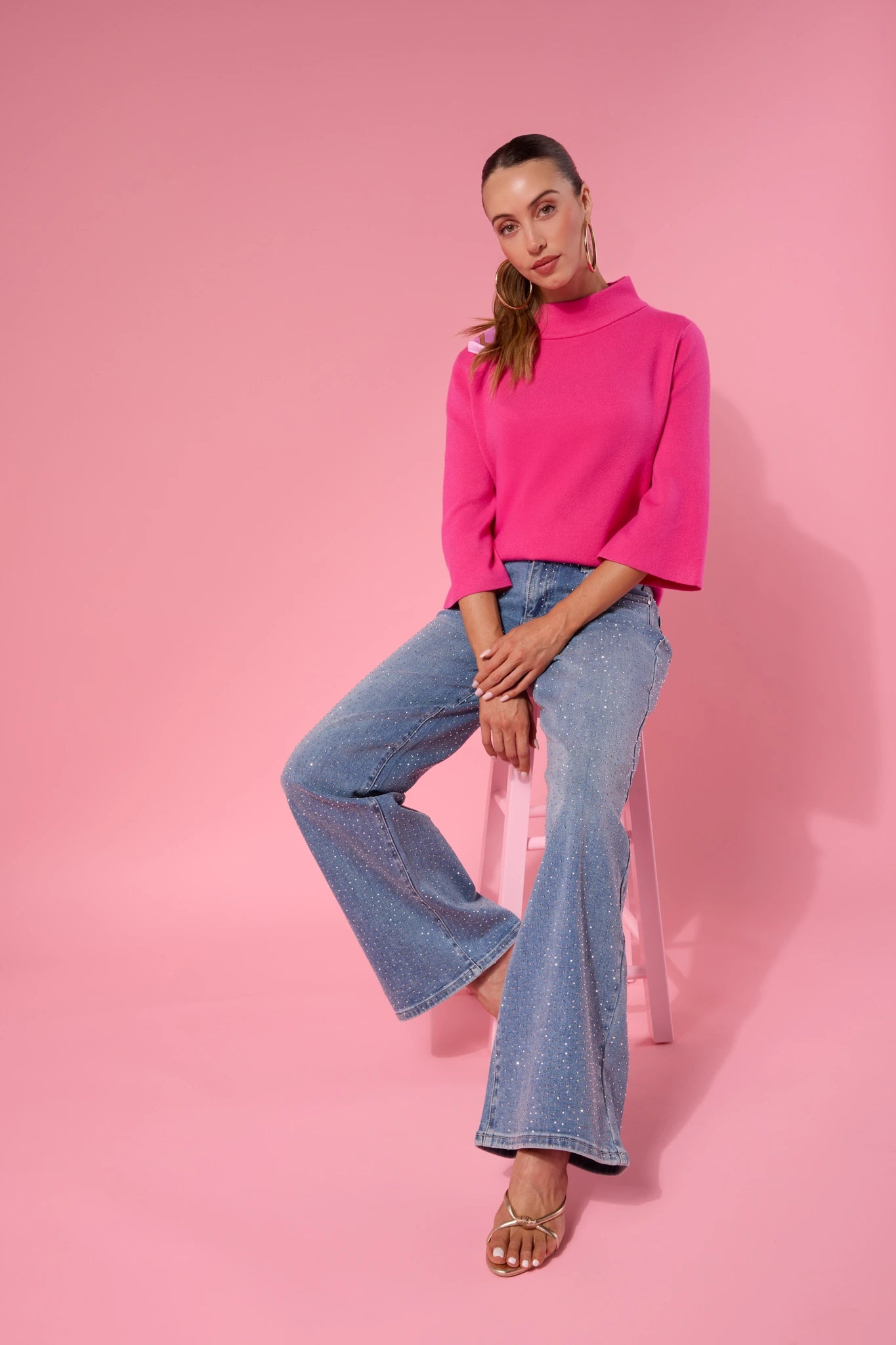 Woman wearing a bright pink Jackie sweater and blue jeans sitting on a pink chair against a pink background