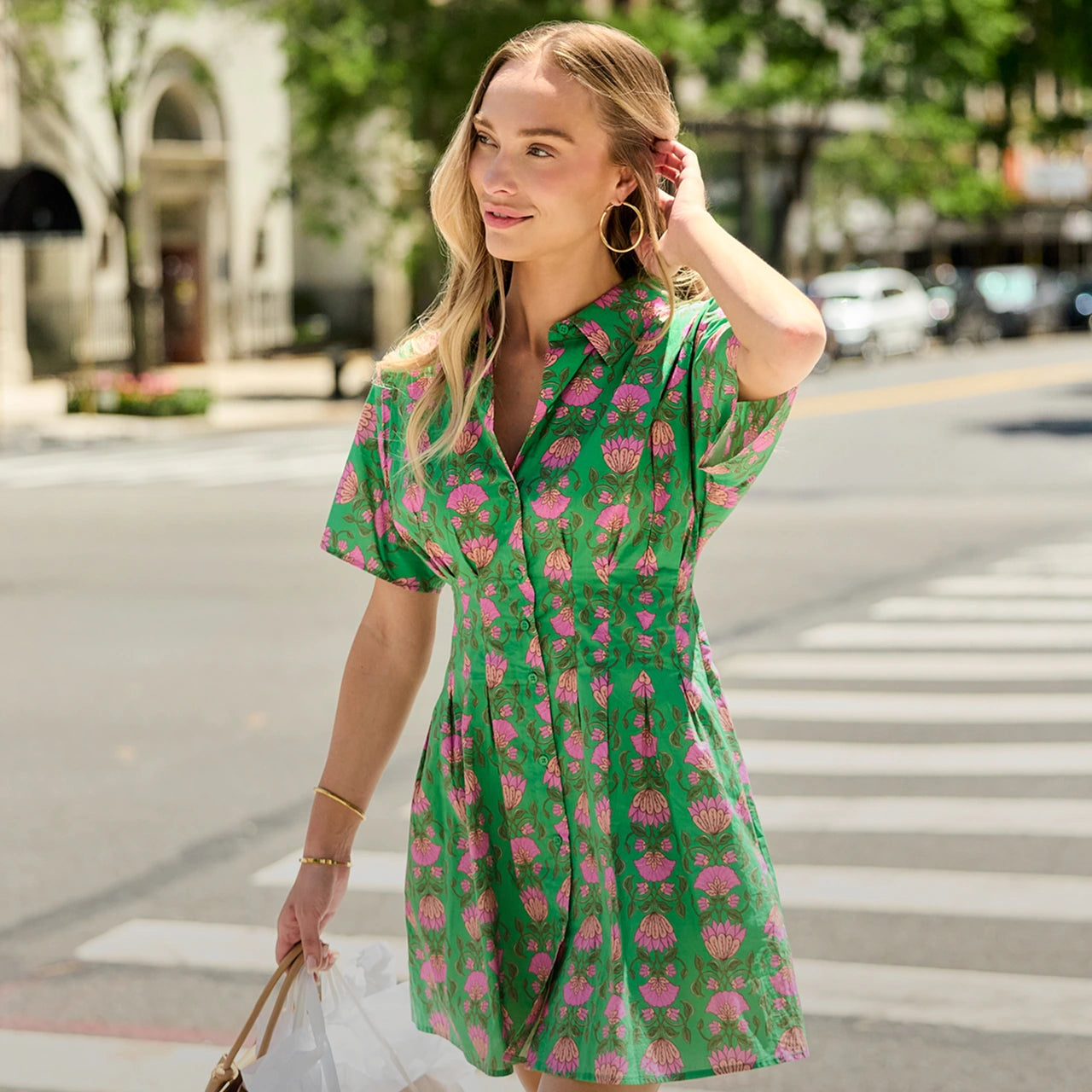 A woman wearing a green short-sleeve collared dress with a pink floral print walks across a city crosswalk. The dress has a button-front neckline and a fitted waist with a flared skirt. hero