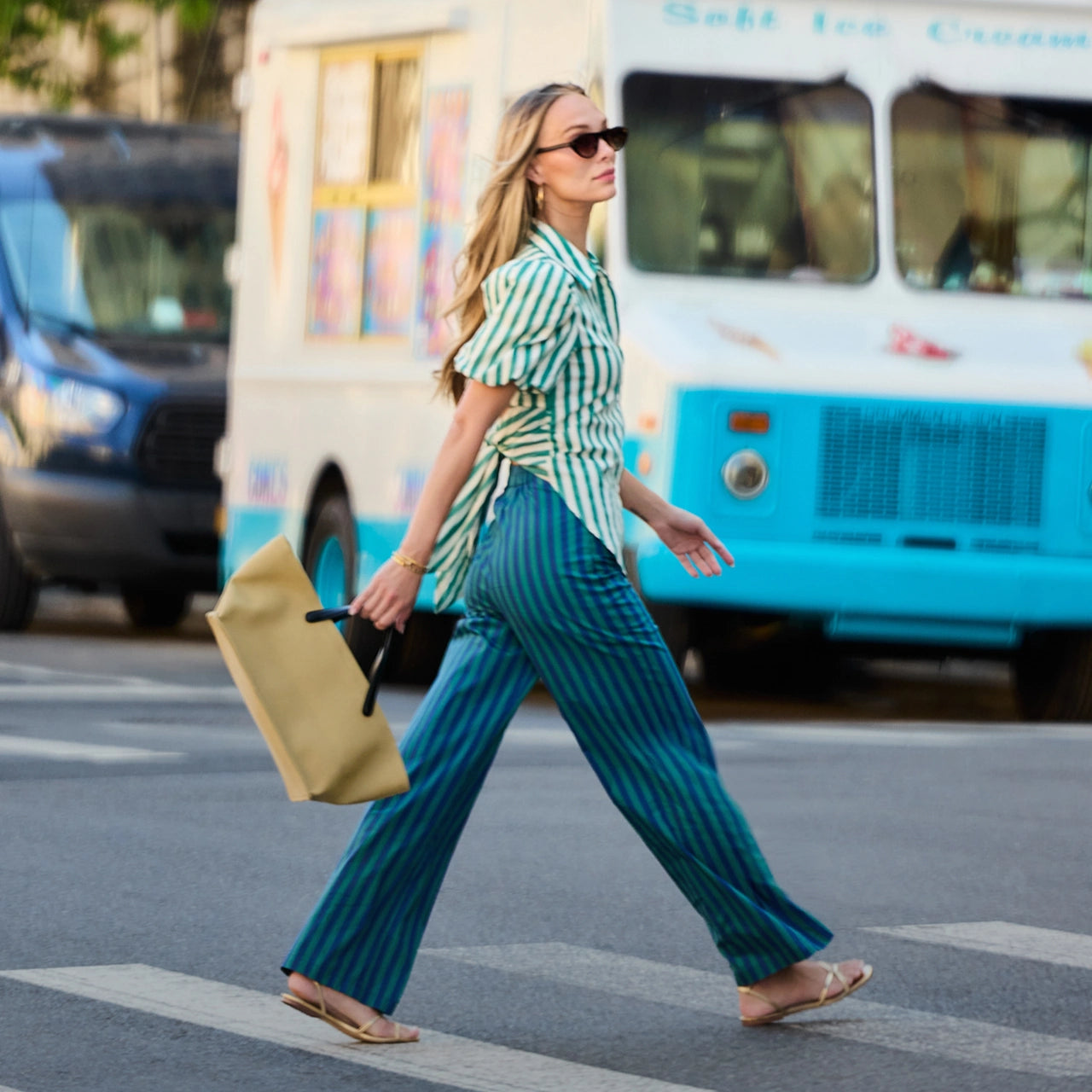 A woman walks across a city crosswalk wearing a green and white striped short-sleeve top and teal pinstriped wide-leg pants, carrying a tan tote bag. A white and blue ice cream truck is blurred in the background. hero
