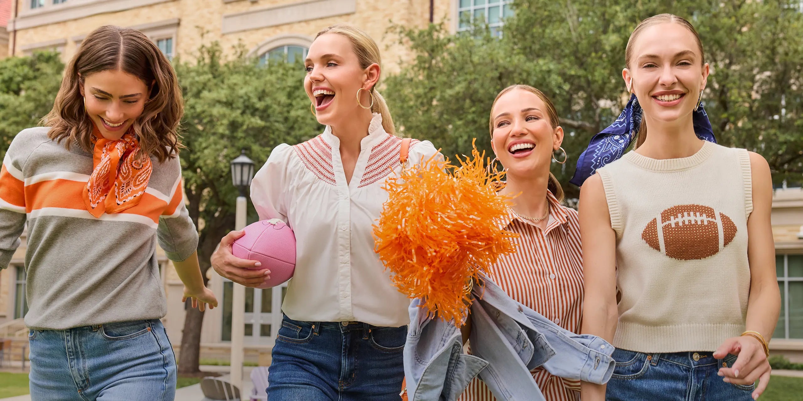 Four women walking outdoors together, smiling and laughing. They are dressed in casual game day outfits, with one holding orange pom-poms and another holding a pink ball