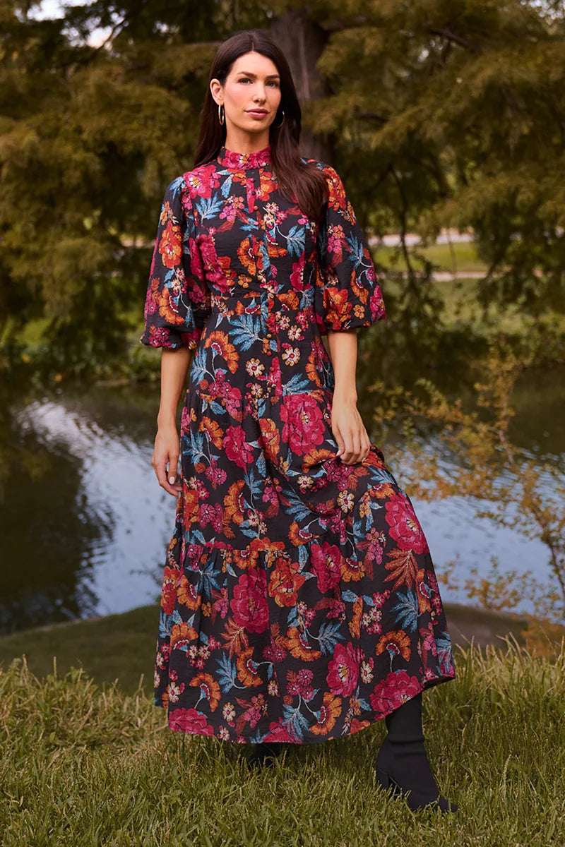Woman wearing a colorful floral dress standing outdoors by a body of water.