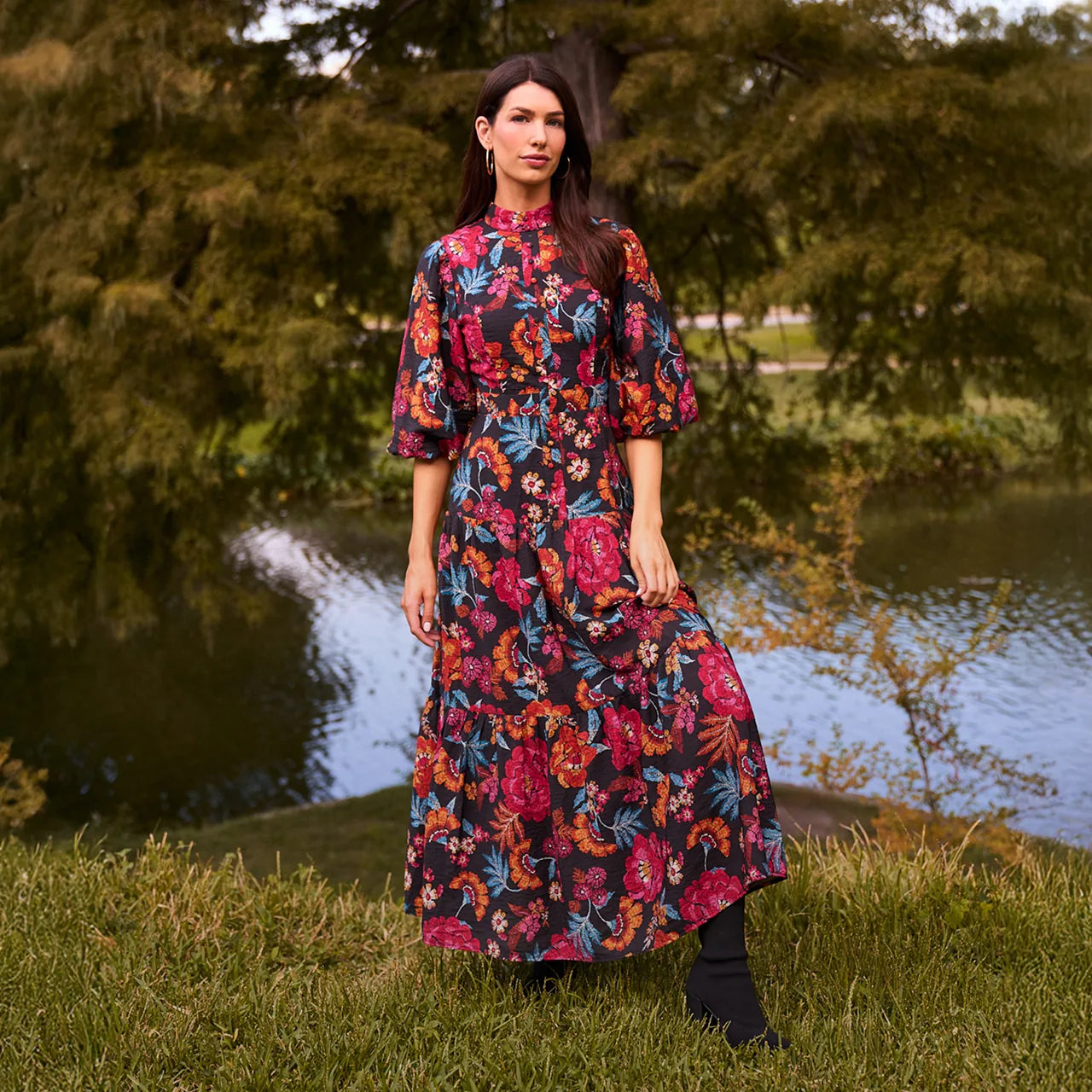 Woman in a colorful floral dress standing by a pond with trees in the background