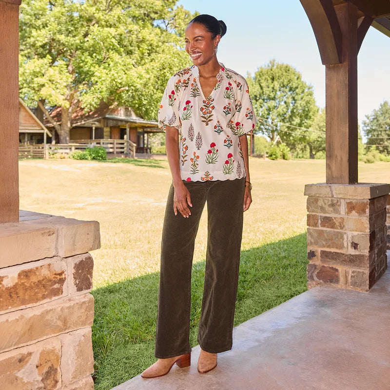 A woman stands on a covered porch wearing a cream short-sleeve blouse with colorful floral embroidery and dark olive wide-leg pants, paired with tan heeled sandals. Trees, grass, and a rustic building are visible in the background.