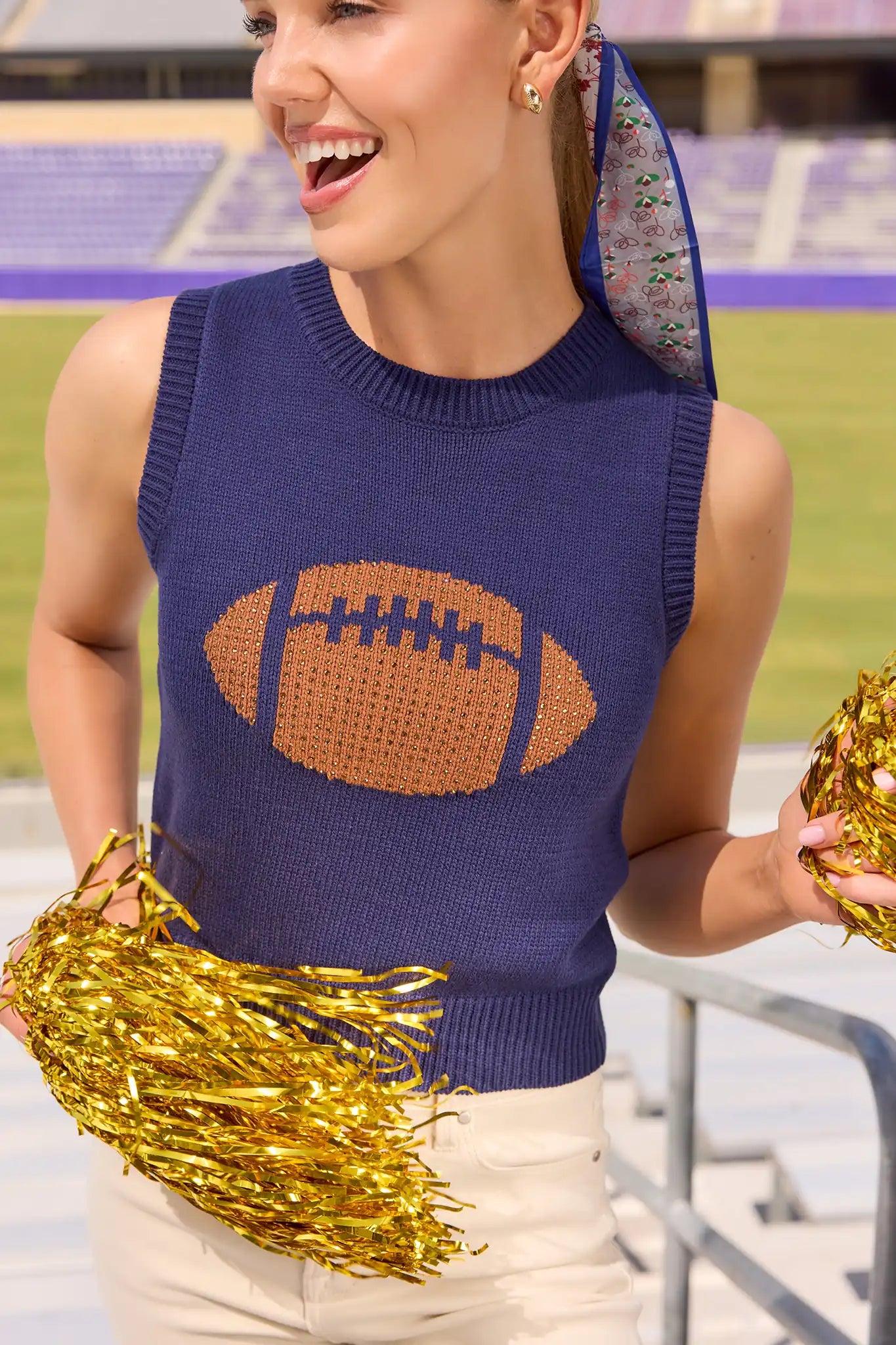 A woman on a football field wearing a navy blue knit sweater with a brown football graphic on the front, holding gold metallic pom-poms.