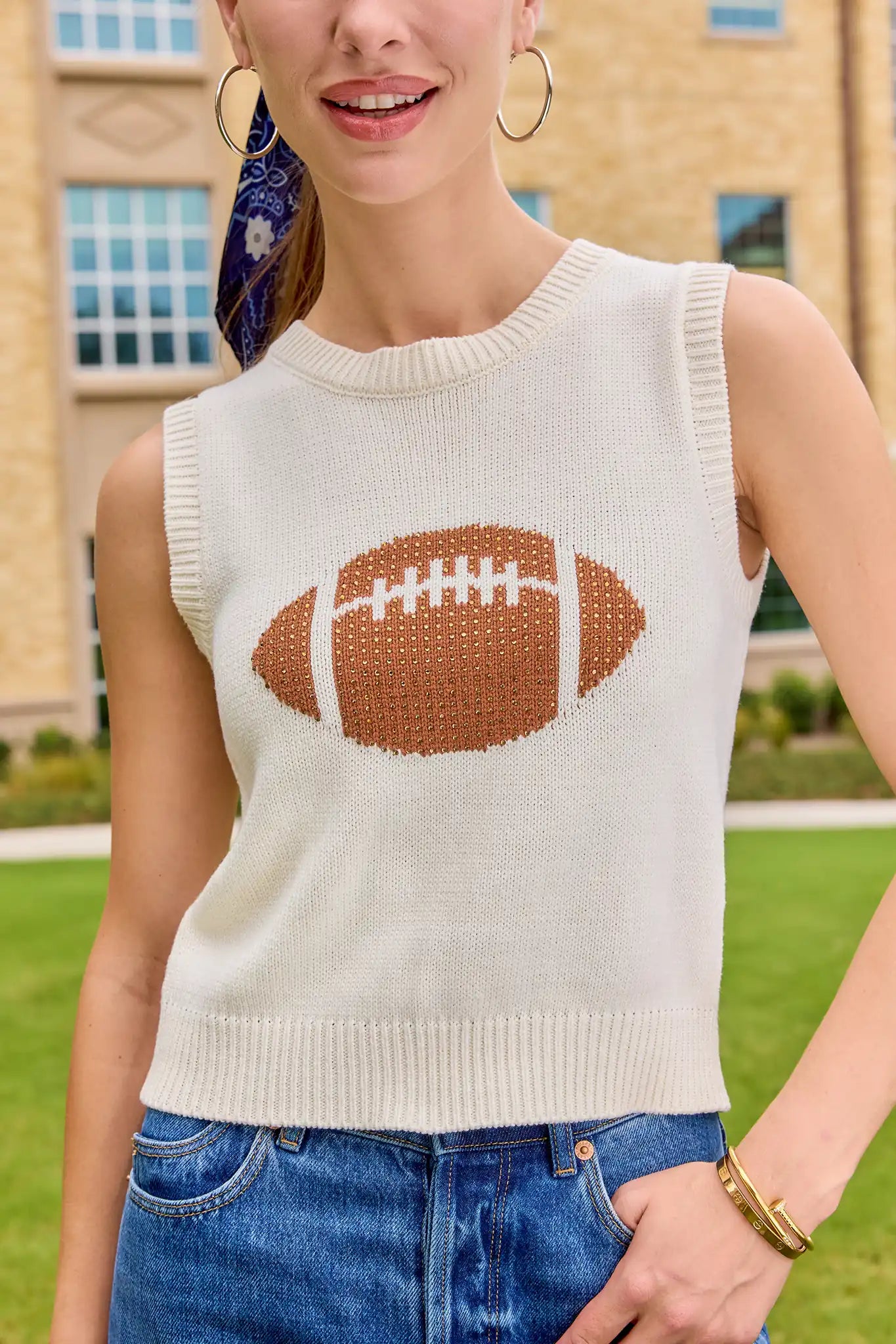 A woman wearing a cream-colored sleeveless knit sweater vest with a brown and white football graphic on the front, paired with blue jeans, standing outdoors in front of a brick building. close 