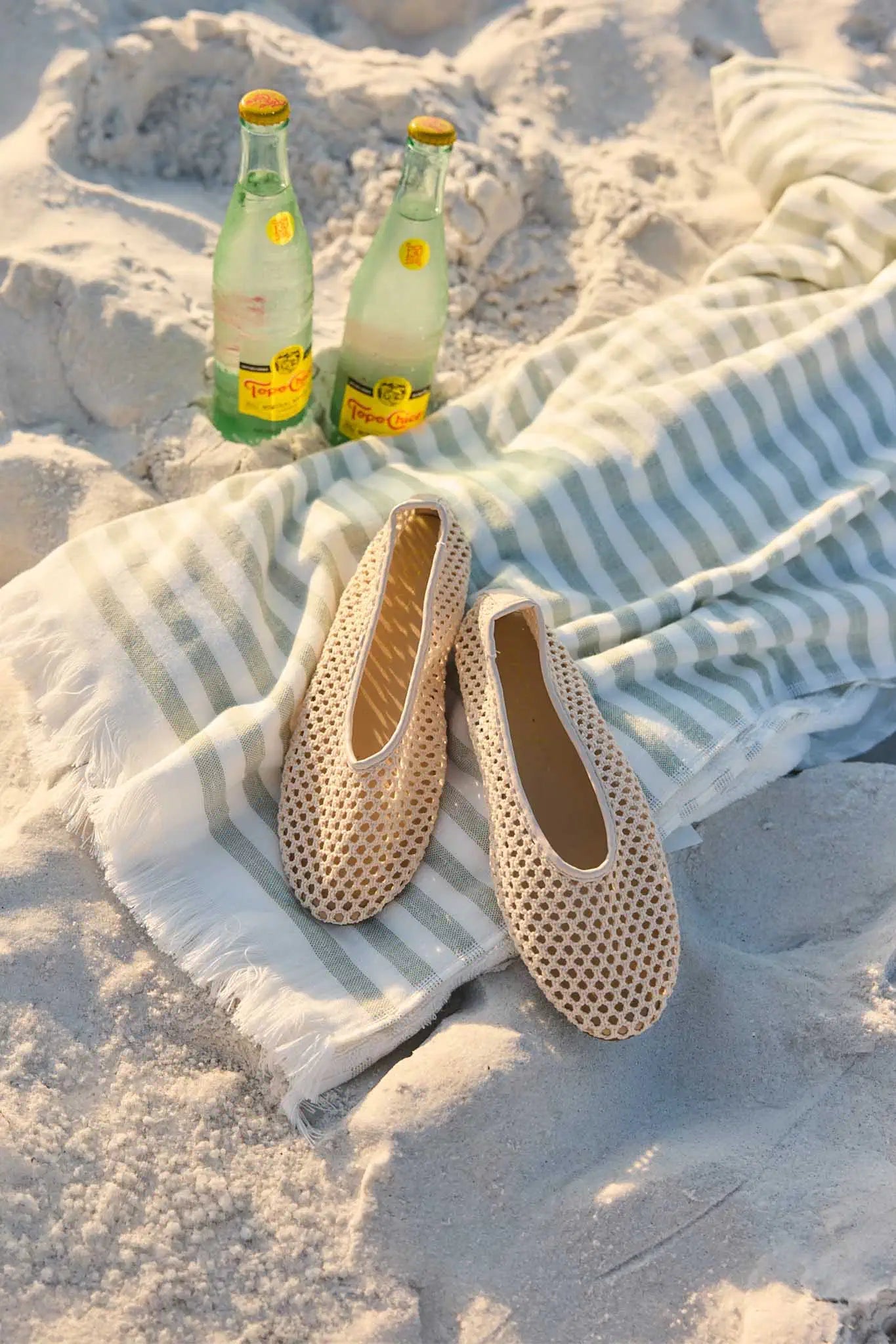 Pair of beige sandals on a striped towel with two bottles in the sand