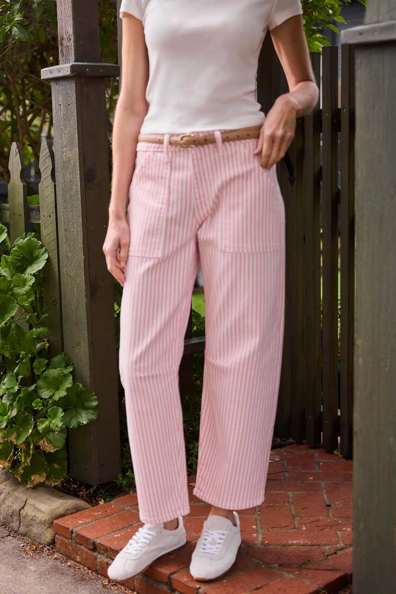 Person wearing pink striped pants and white shoes standing on a brick path with greenery in the background