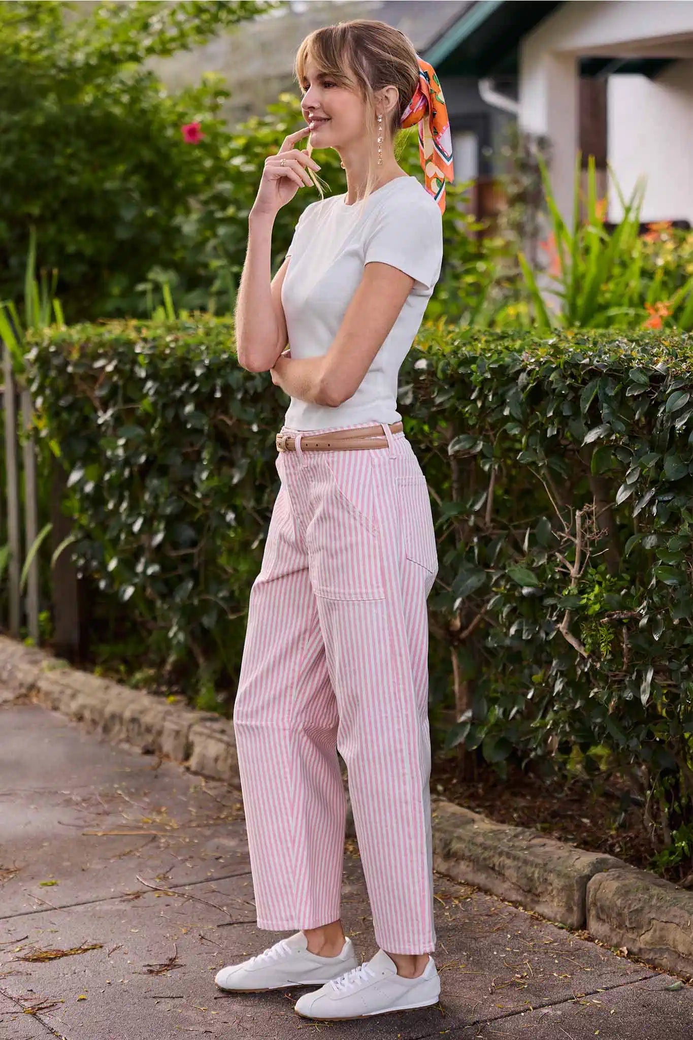 Woman in white top and pink striped pants standing outdoors with greenery in the background