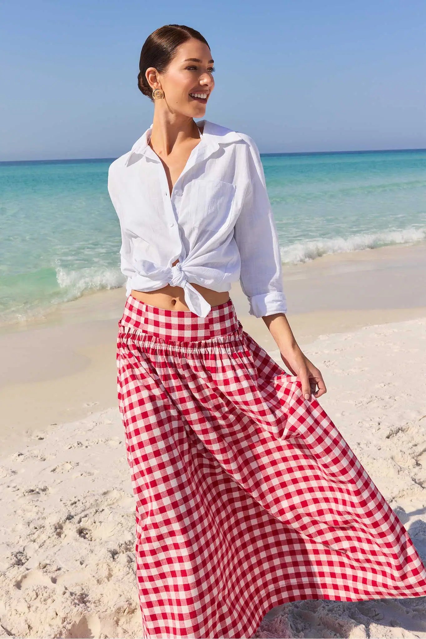 Woman in white shirt and red checkered pants standing on a beach with clear blue water.