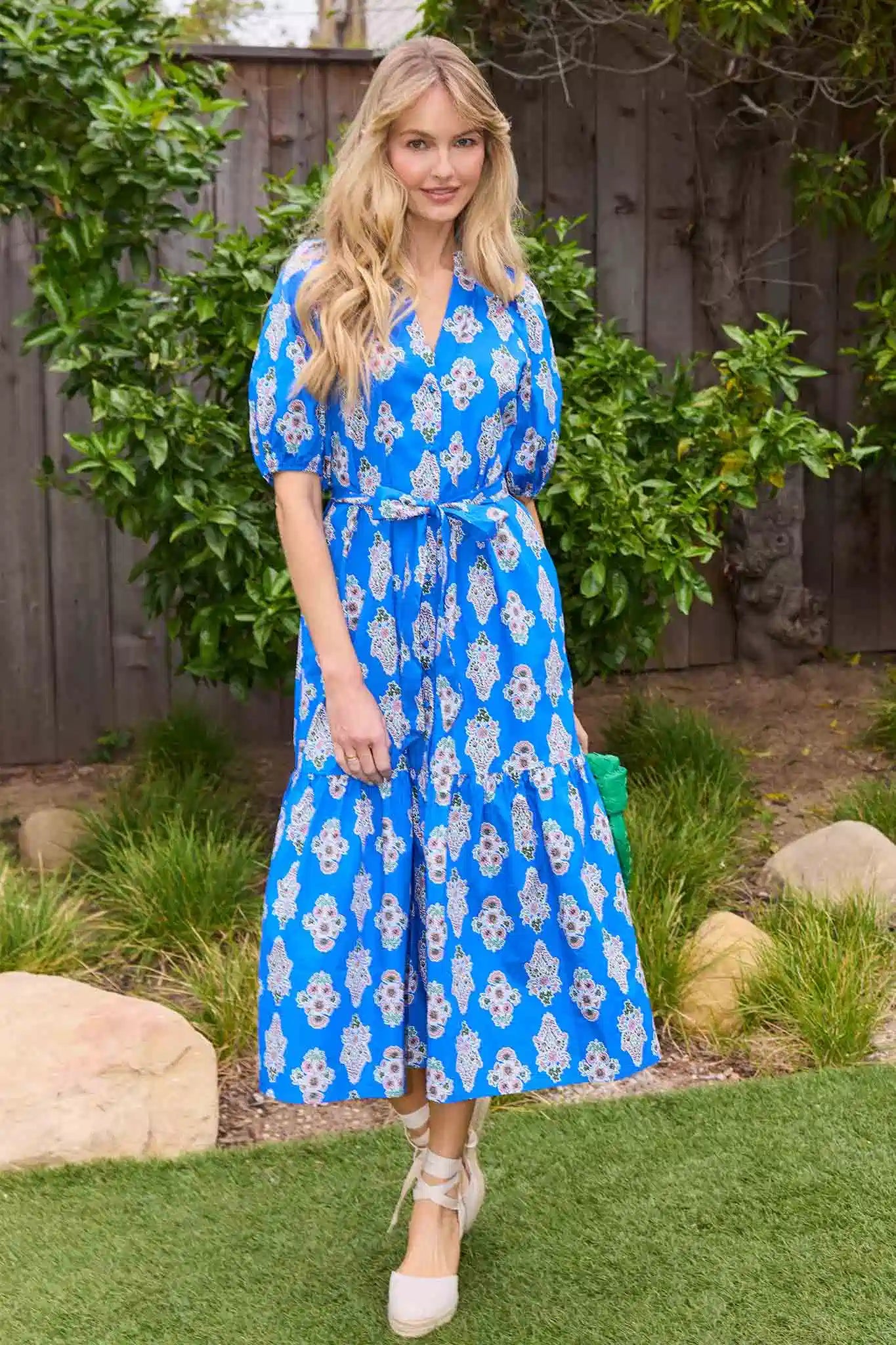 Woman wearing a blue floral dress standing outdoors with greenery in the background