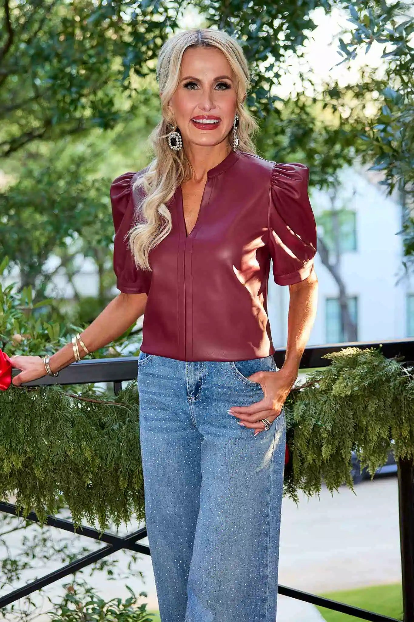 Woman wearing a burgundy leather Ariana Top and blue jeans standing outdoors with greenery in the background