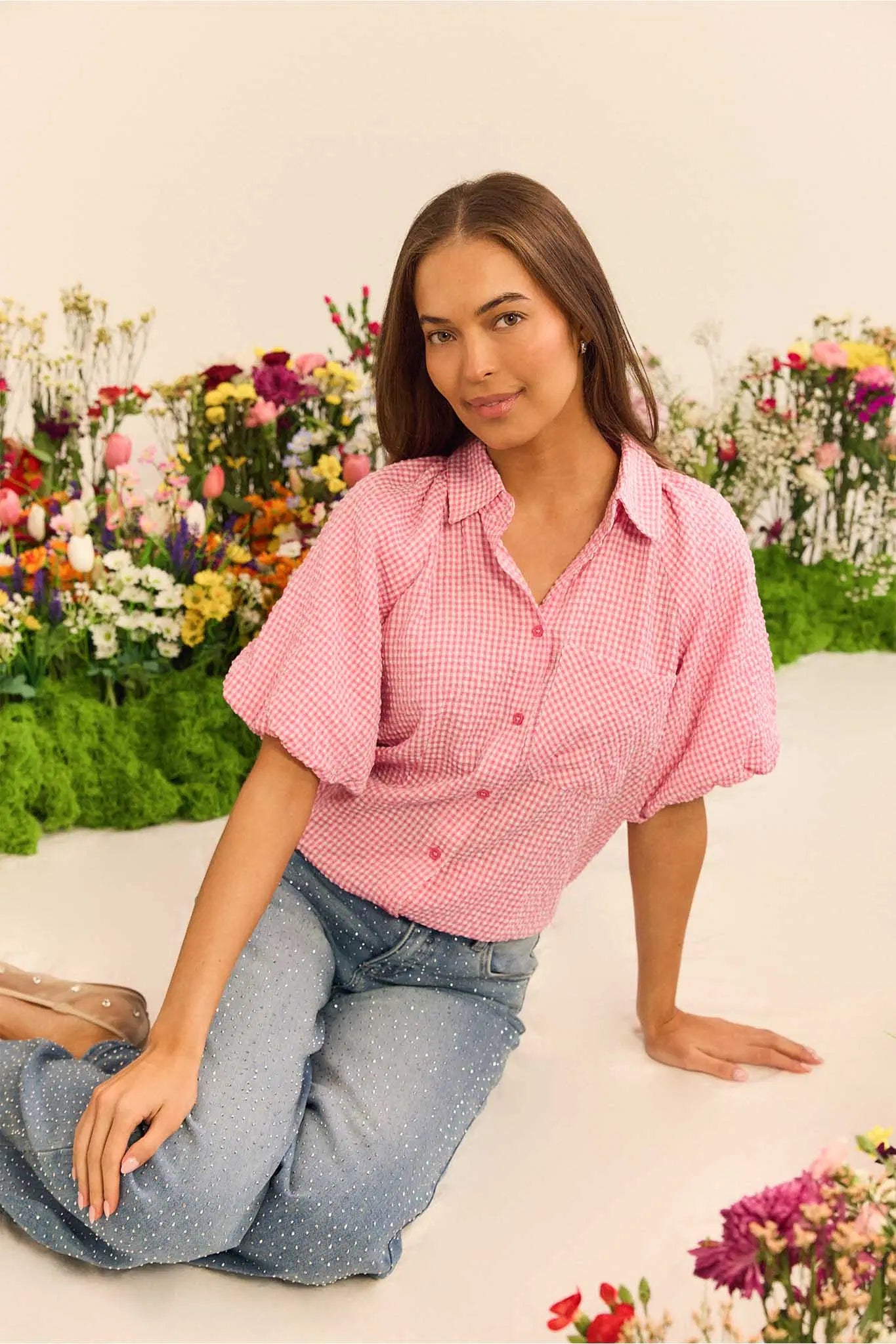 Woman wearing a pink checkered shirt and blue jeans sitting on a white surface with floral decorations in the background.