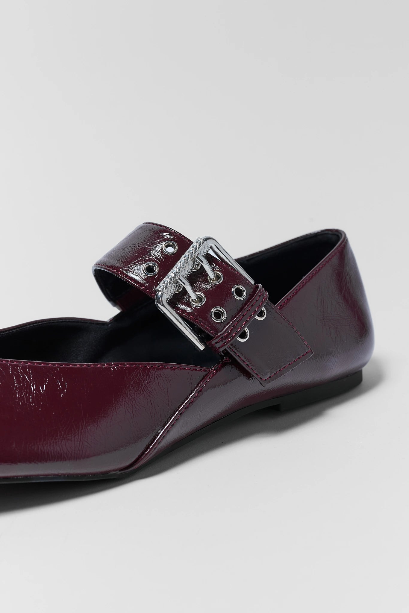 A close-up of a glossy burgundy faux leather flat shoe featuring a wide strap with silver-tone metal eyelets and a decorative silver buckle.