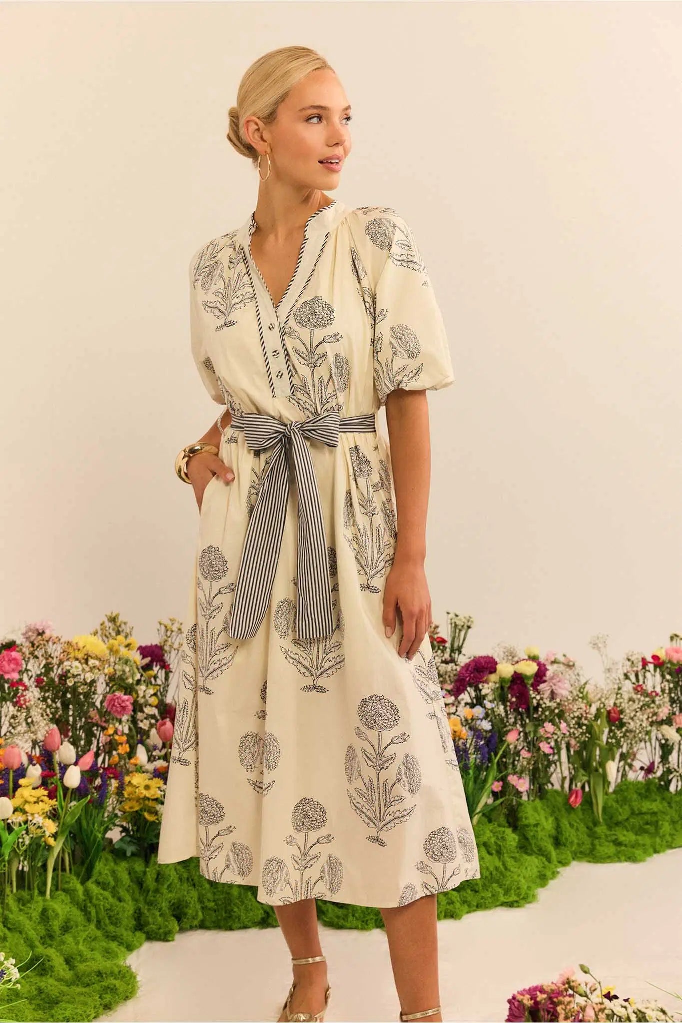 Woman wearing a floral dress with a belt, standing in front of a floral garden.