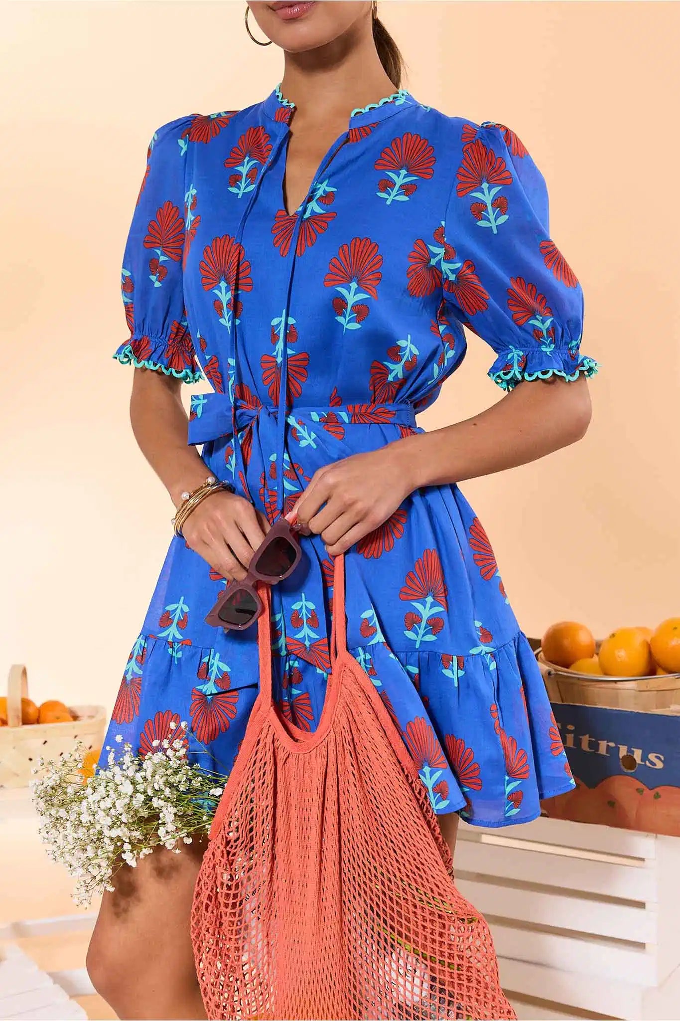 Woman wearing a blue dress with red and green patterns, holding an orange bag against a beige background.