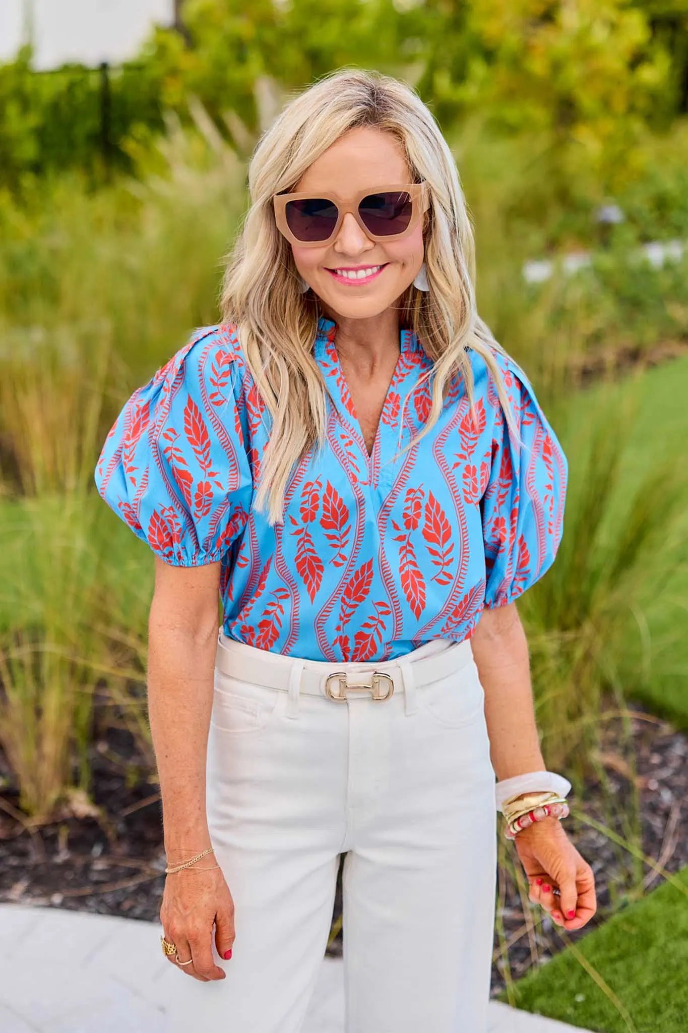 Woman wearing a blue patterned blouse and white pants outdoors.