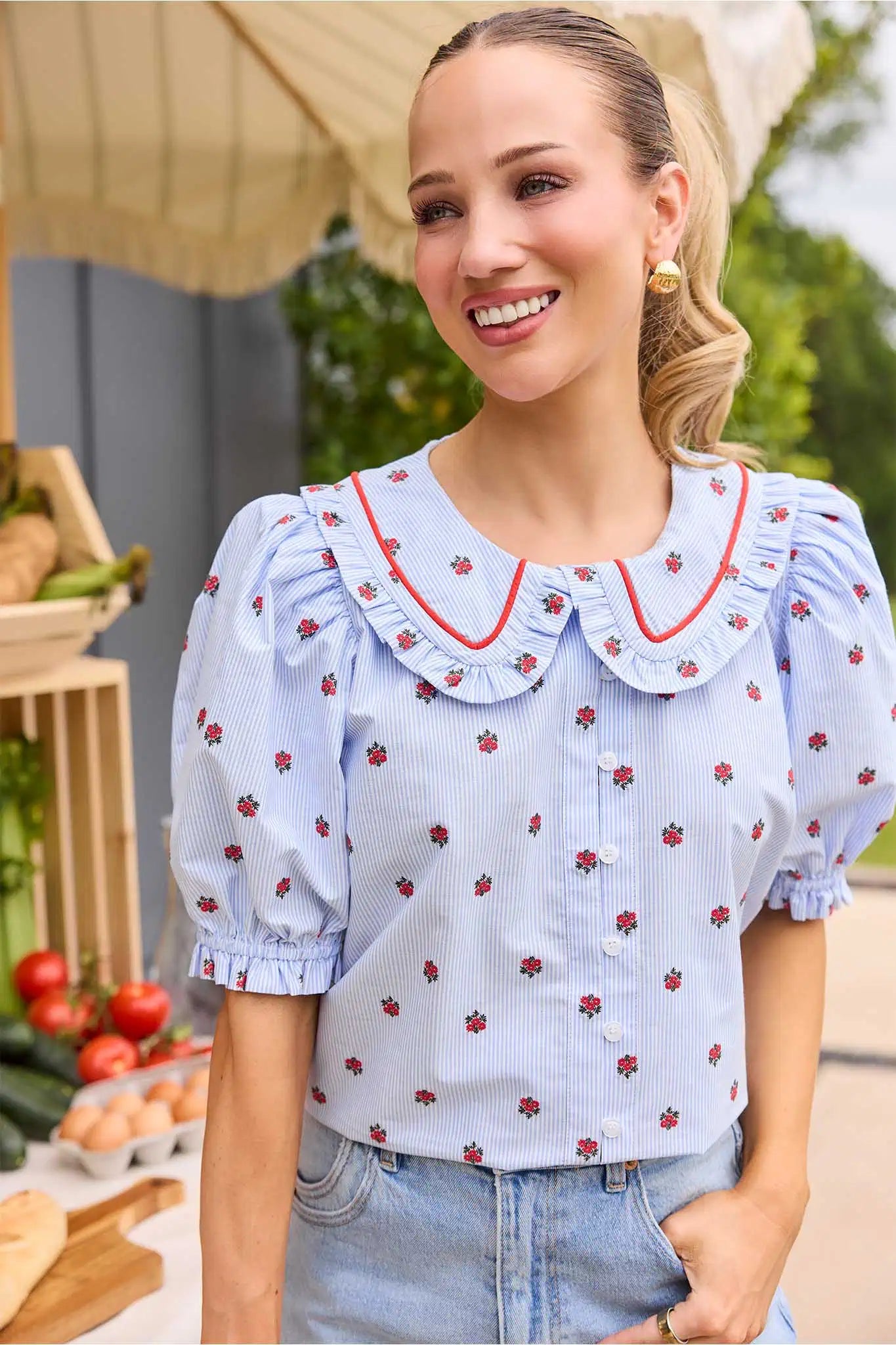 Woman wearing a light blue blouse with red floral pattern and ruffled collar, standing outdoors.